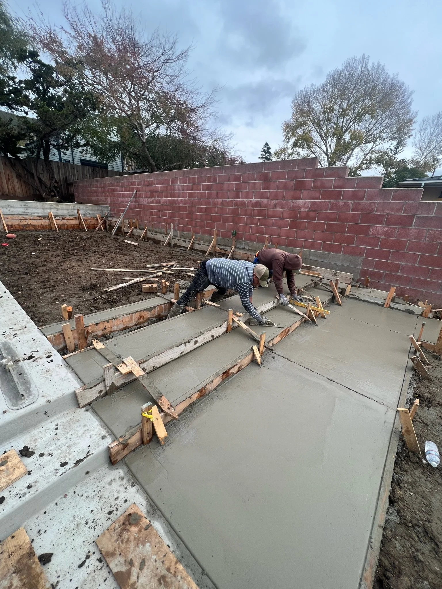 Two workers are pouring and smoothing concrete for a sidewalk or patio, with wooden forms and stakes in place. The background features a brick wall and trees under a cloudy sky.