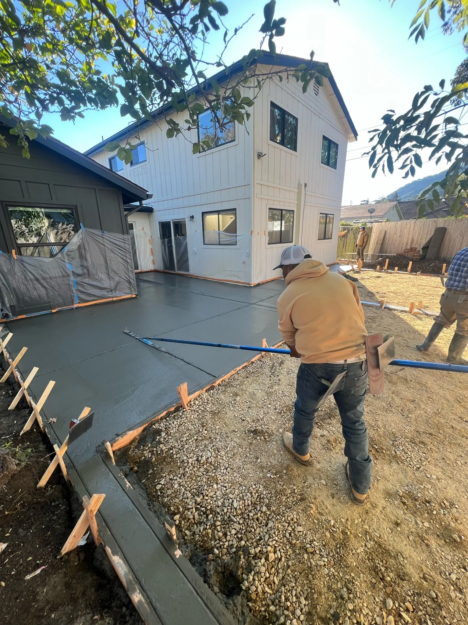 Workers pouring wet concrete for a new patio in the backyard of a two-story house, with wooden framing and construction tools visible.