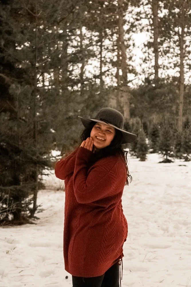 A smiling woman wearing a wide-brimmed hat and a red sweater standing in a snowy forest.