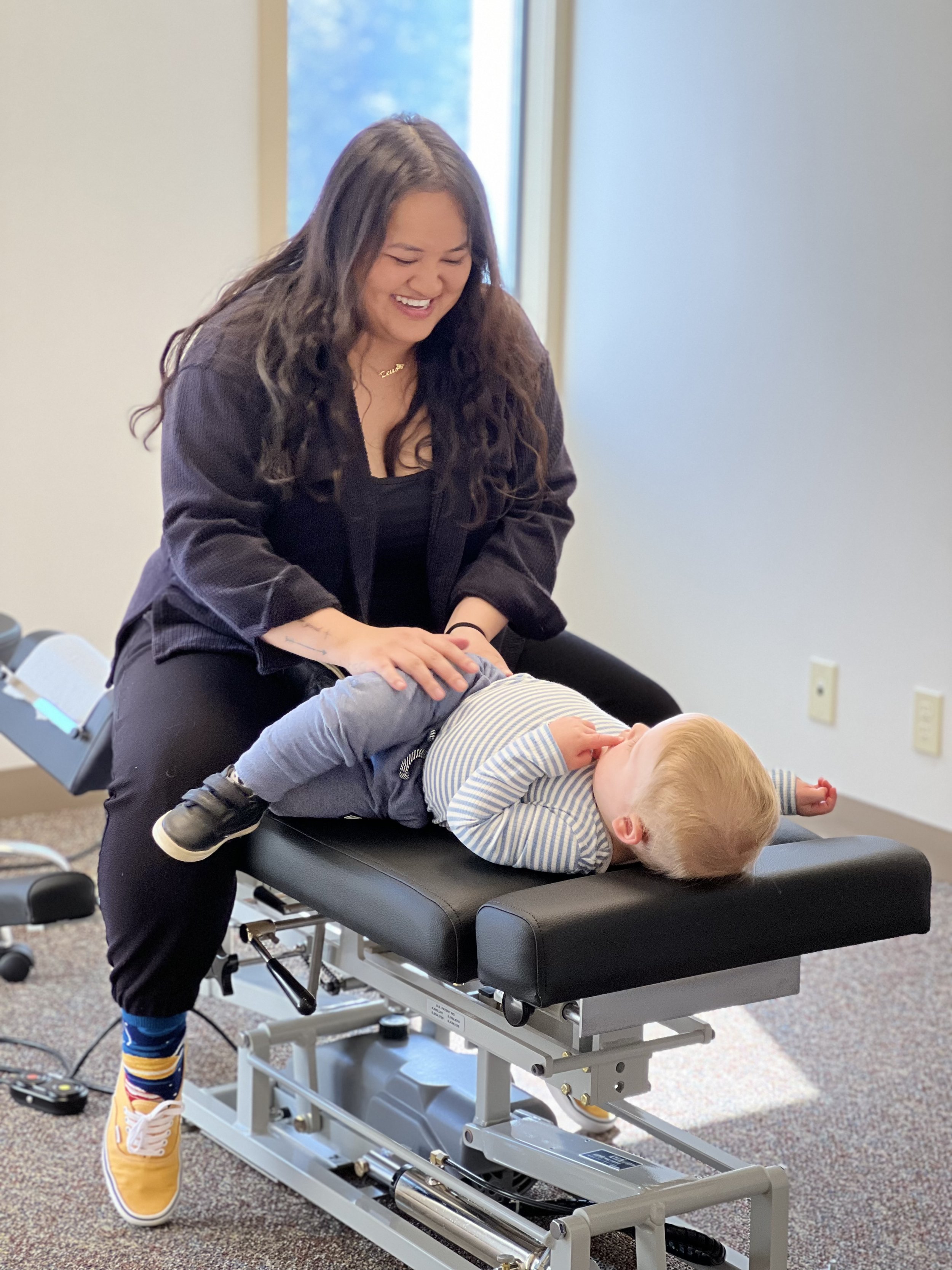 A woman with long dark hair laughing as she holds a young blonde boy lying face down on a chiropractic examination table in a clinic. The woman is wearing a black jacket and colorful striped socks with yellow sneakers.