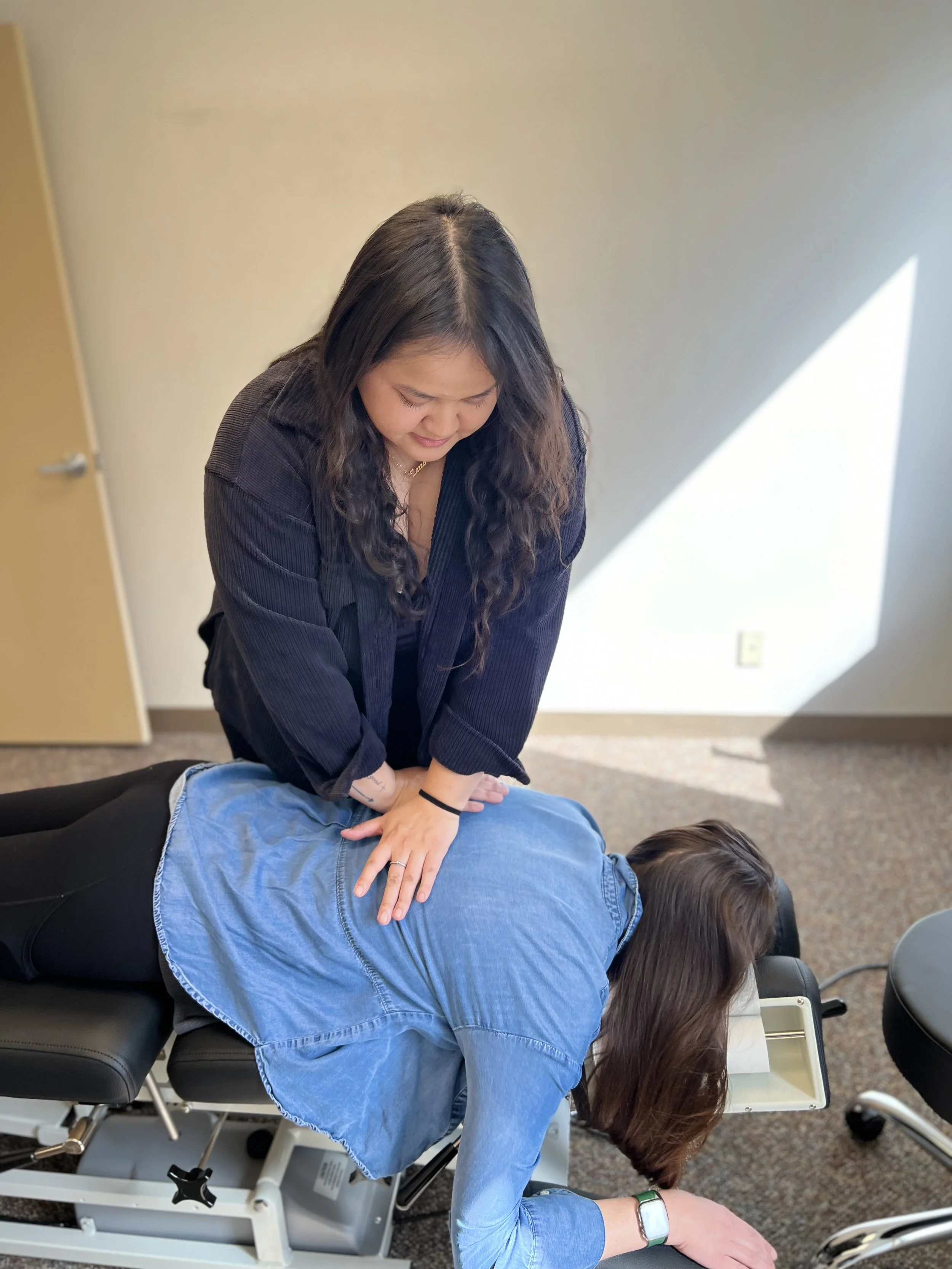 A woman performing a chiropractic adjustment on a person lying face down on a therapy table in an office.