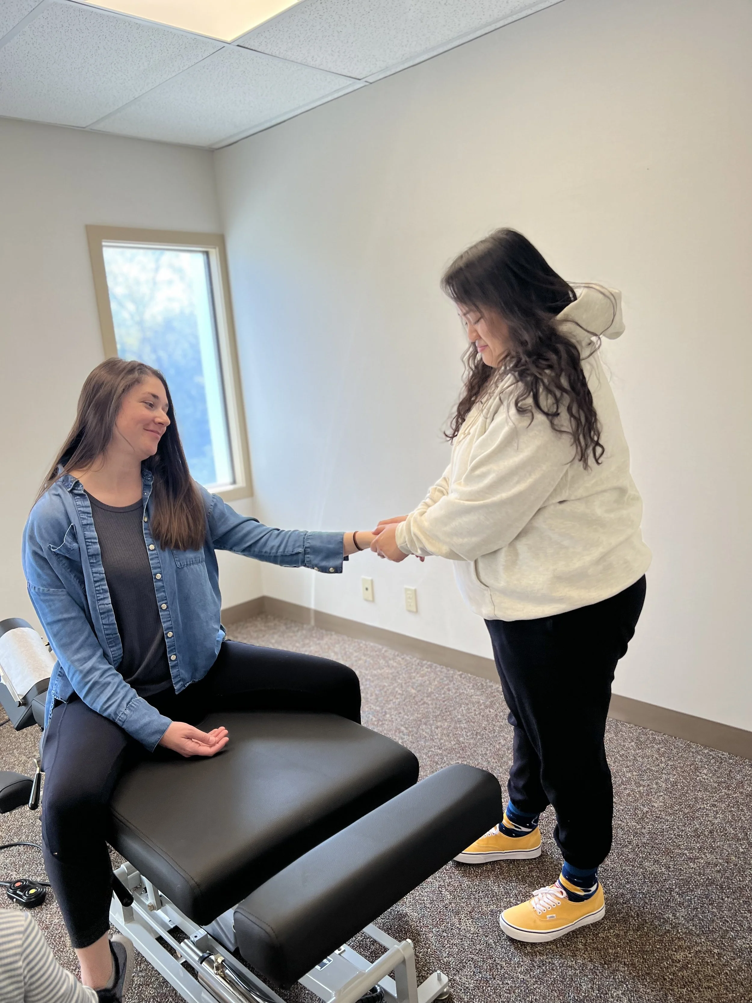 A woman is sitting on a black massage table with her eyes closed and hands resting on her lap. Another woman, standing and smiling, is holding her hands and leaning slightly forward. The room has white walls, a window with natural light, and a carpeted floor. The standing woman is wearing a beige hoodie, black pants, and yellow sneakers.