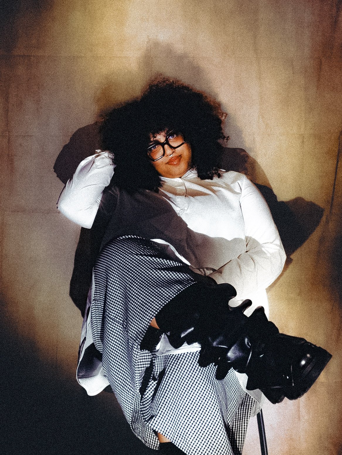 A woman with curly black hair, glasses, and wearing a white shirt, sitting in a chair against a plain background, looking at the camera with a slight smile.