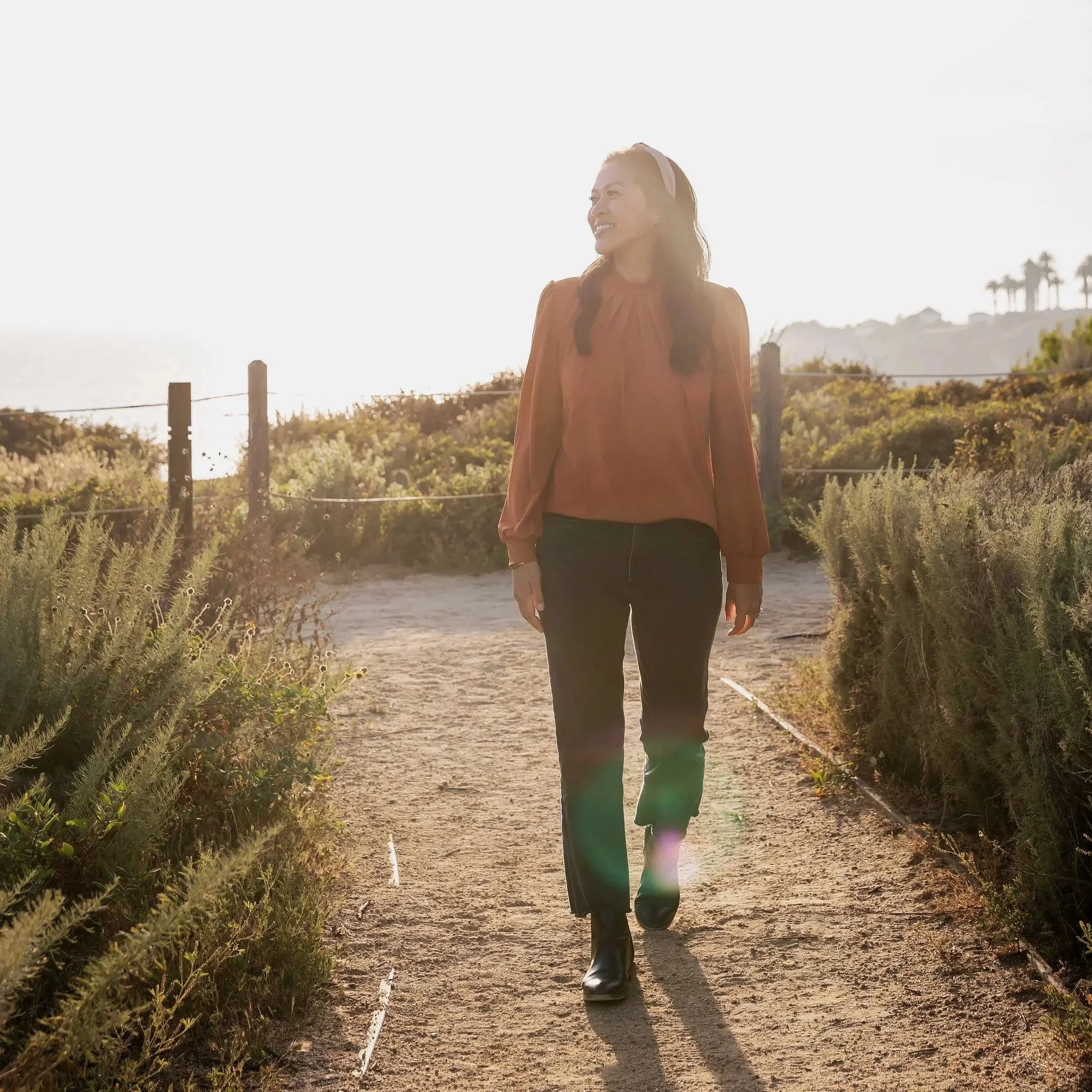 A woman walking outdoors on a dirt path, wearing a rust-colored blouse, demin jeans, and black boots, smiling, with sunlight behind her and some houses and palm trees in the distance.