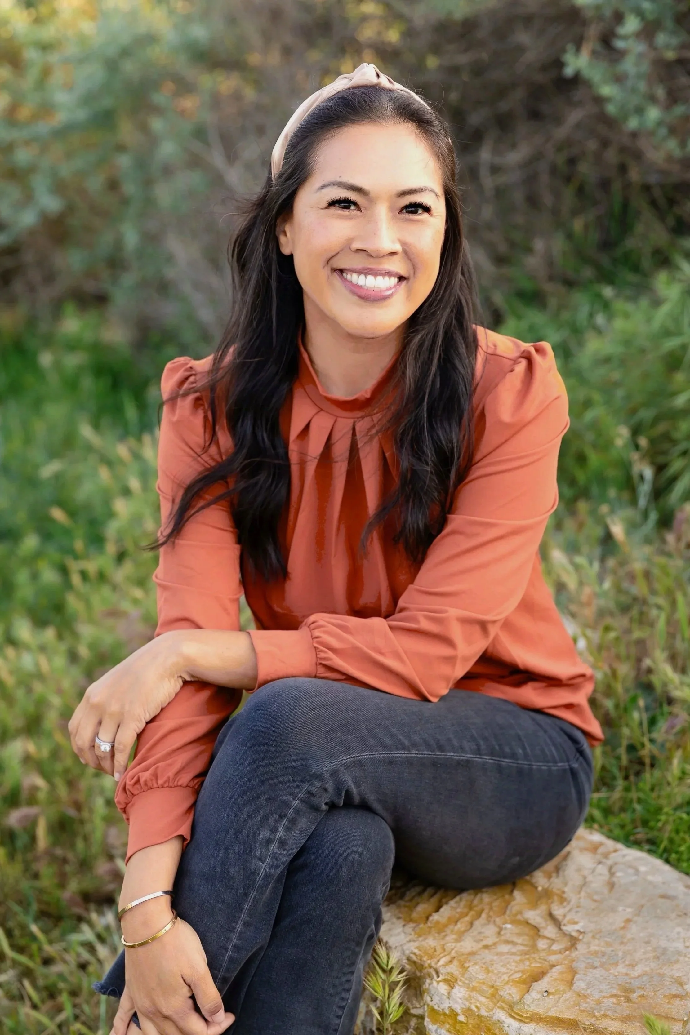 A woman with long dark hair, wearing an rust-colored blouse and dark jeans, smiling and sitting on a log outdoors with greenery in the background.