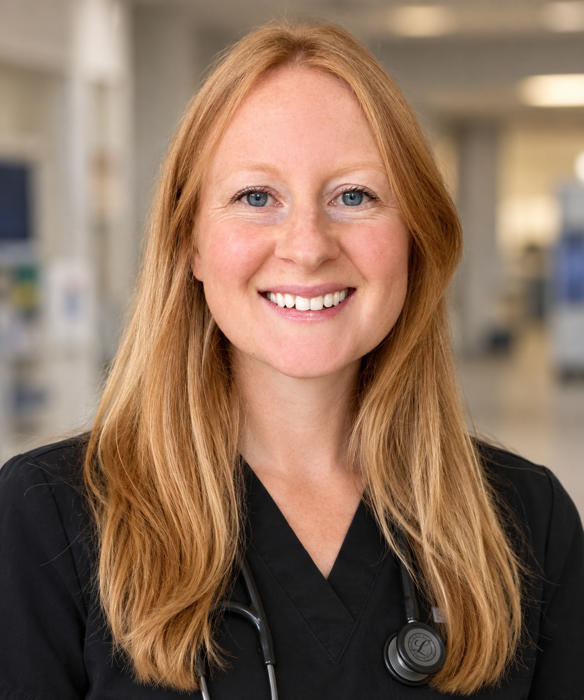 Photograph of a smiling female healthcare professional with long red hair, wearing black scrubs and a stethoscope around her neck, in a medical facility.