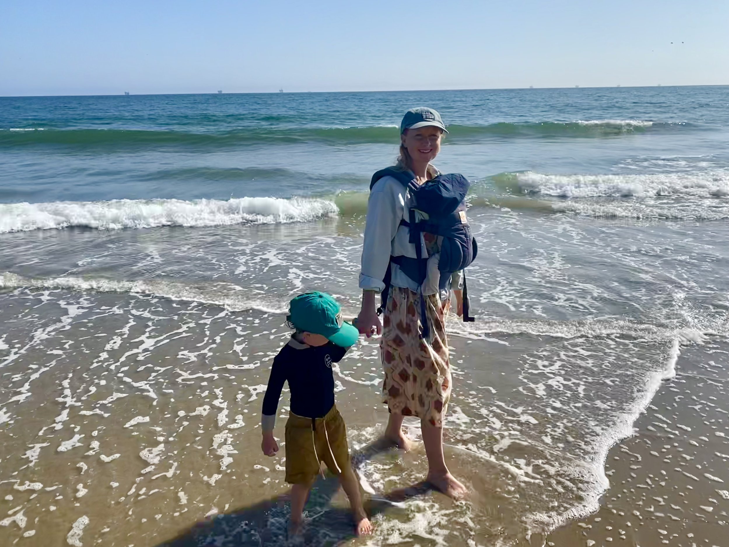 A woman and a child walking on the beach with waves and ocean behind them. The woman is smiling, wearing a hat, a light blue jacket, and a patterned skirt. The child is wearing a teal hat, a dark long-sleeve shirt, and brown shorts.