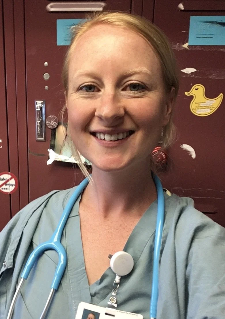 A smiling female healthcare worker in gray scrubs with a blue stethoscope around her neck, standing in front of maroon lockers decorated with duck stickers and feathers.