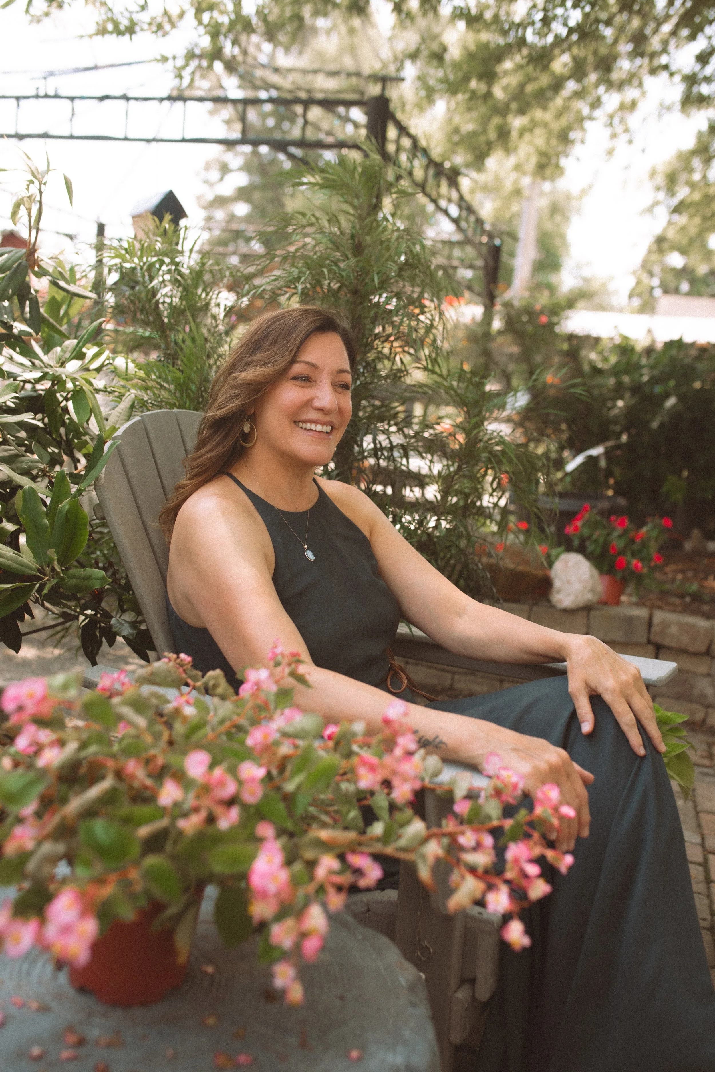 A woman with brown hair, earrings, and a black sleeveless top sitting on a garden chair outdoors, smiling, surrounded by plants and flowers.