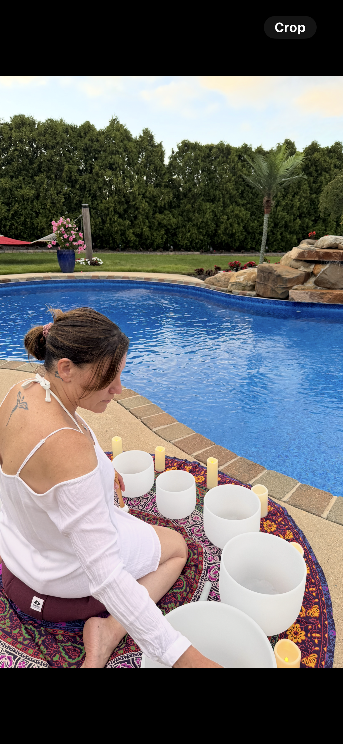 Woman in white shirt sitting on patterned blanket by swimming pool with singing bowls and candles for a meditation session in backyard.