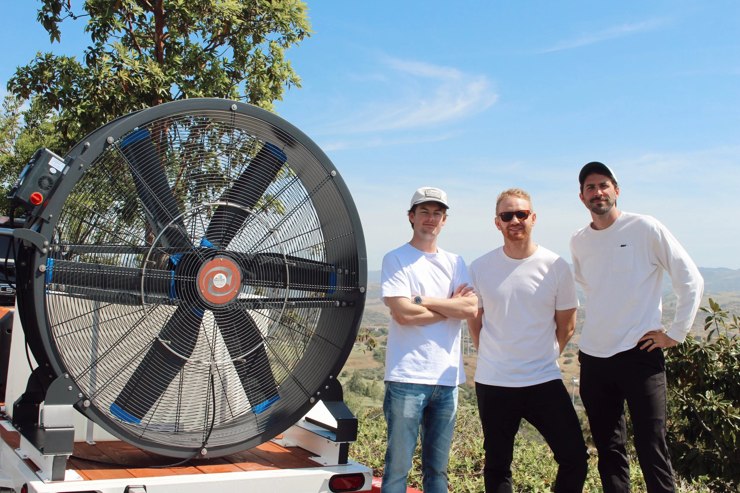 Three men standing outdoors in front of a large cooling fan or industrial fan, with clear blue skies and trees in the background.