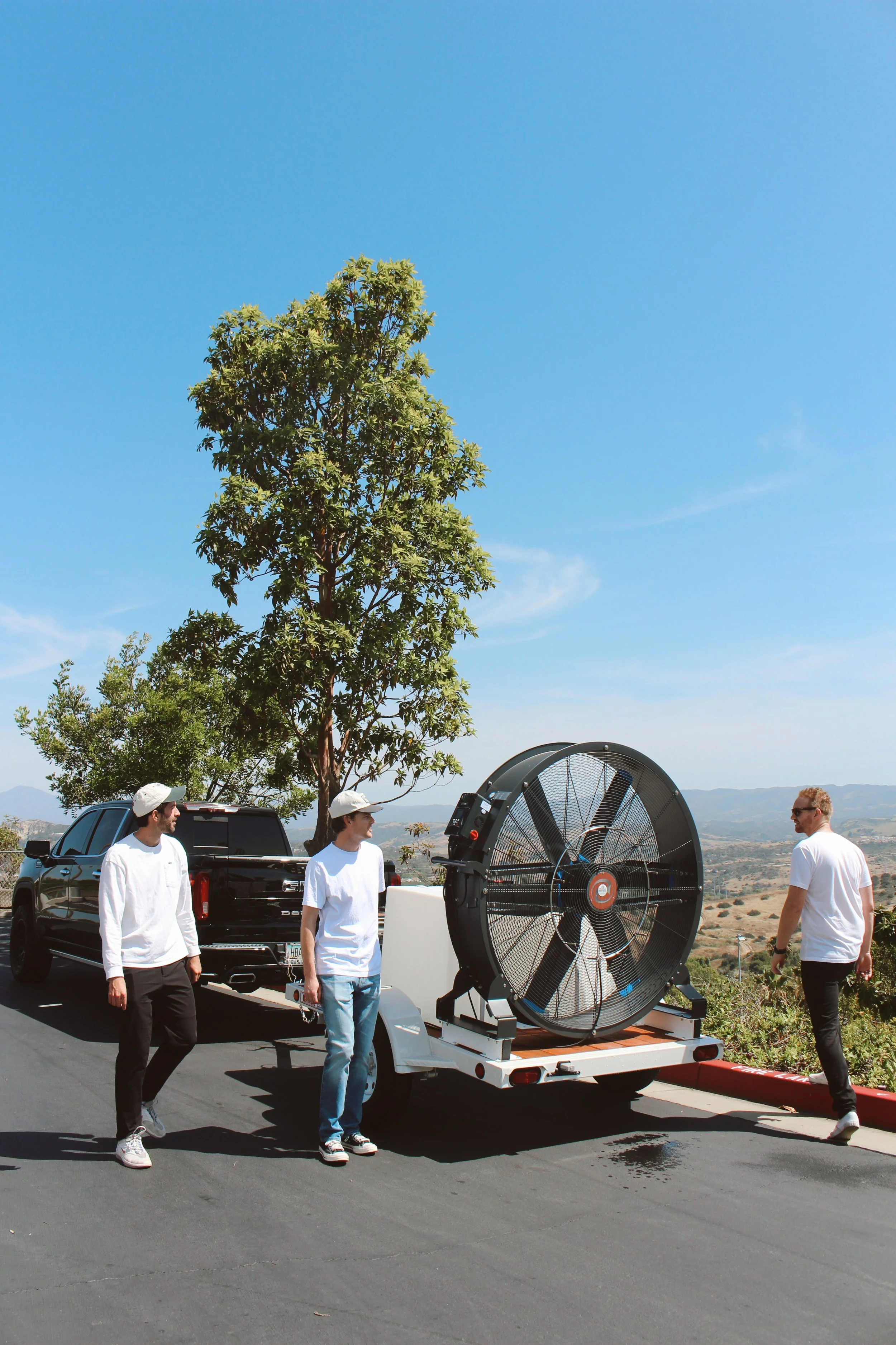 Four men in white shirts and hats standing near a large industrial fan mounted on a trailer, with a black pickup truck in the background, outdoors on a clear day with blue skies and rolling hills.