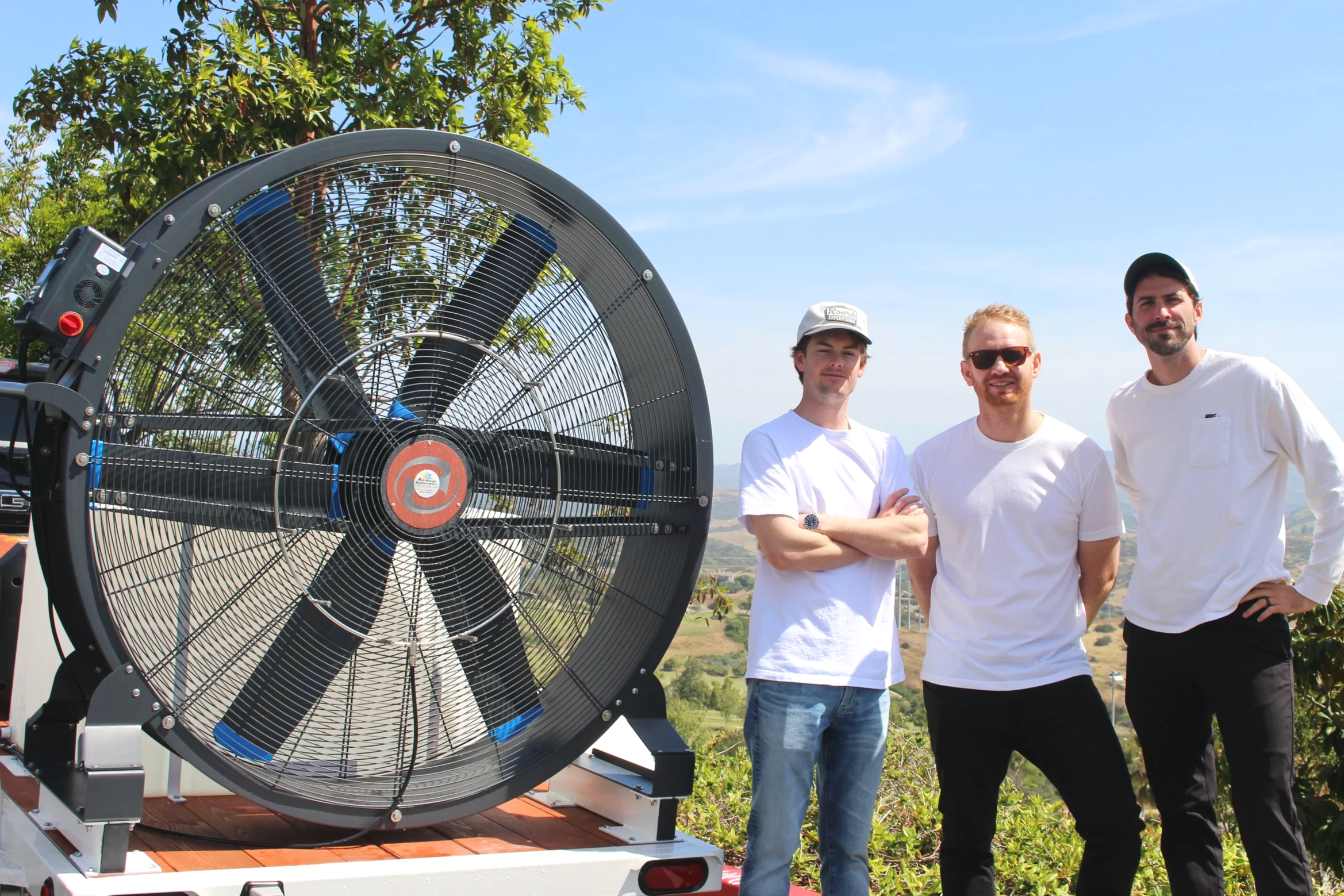 Three men stand outdoors next to a large industrial wind turbine fan. One man has crossed arms, another has hands on hips, and the third has hands by his sides. The background features trees and a clear blue sky.