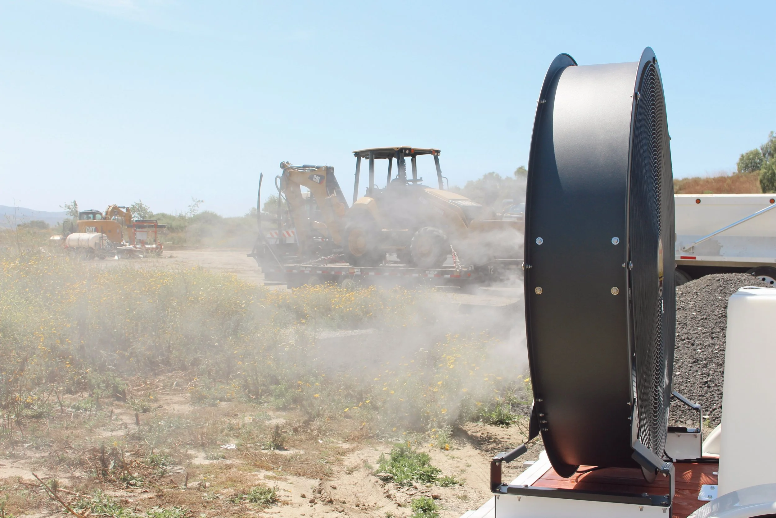 A dust suppression system in operation near construction equipment on a dry, grassy field under a clear blue sky.