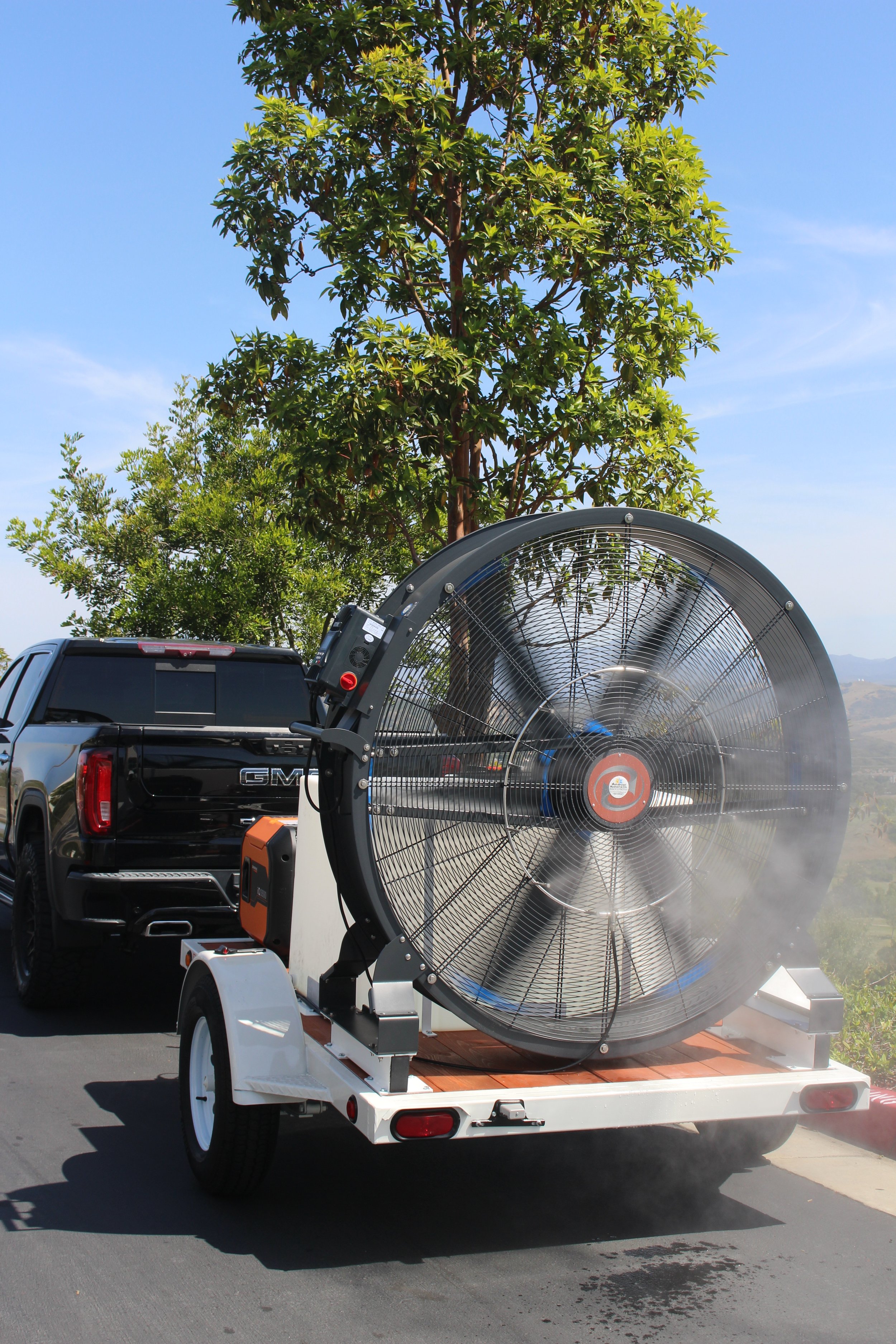 A large industrial fan mounted on a trailer, pulled by a black pickup truck, with trees and a clear blue sky in the background.