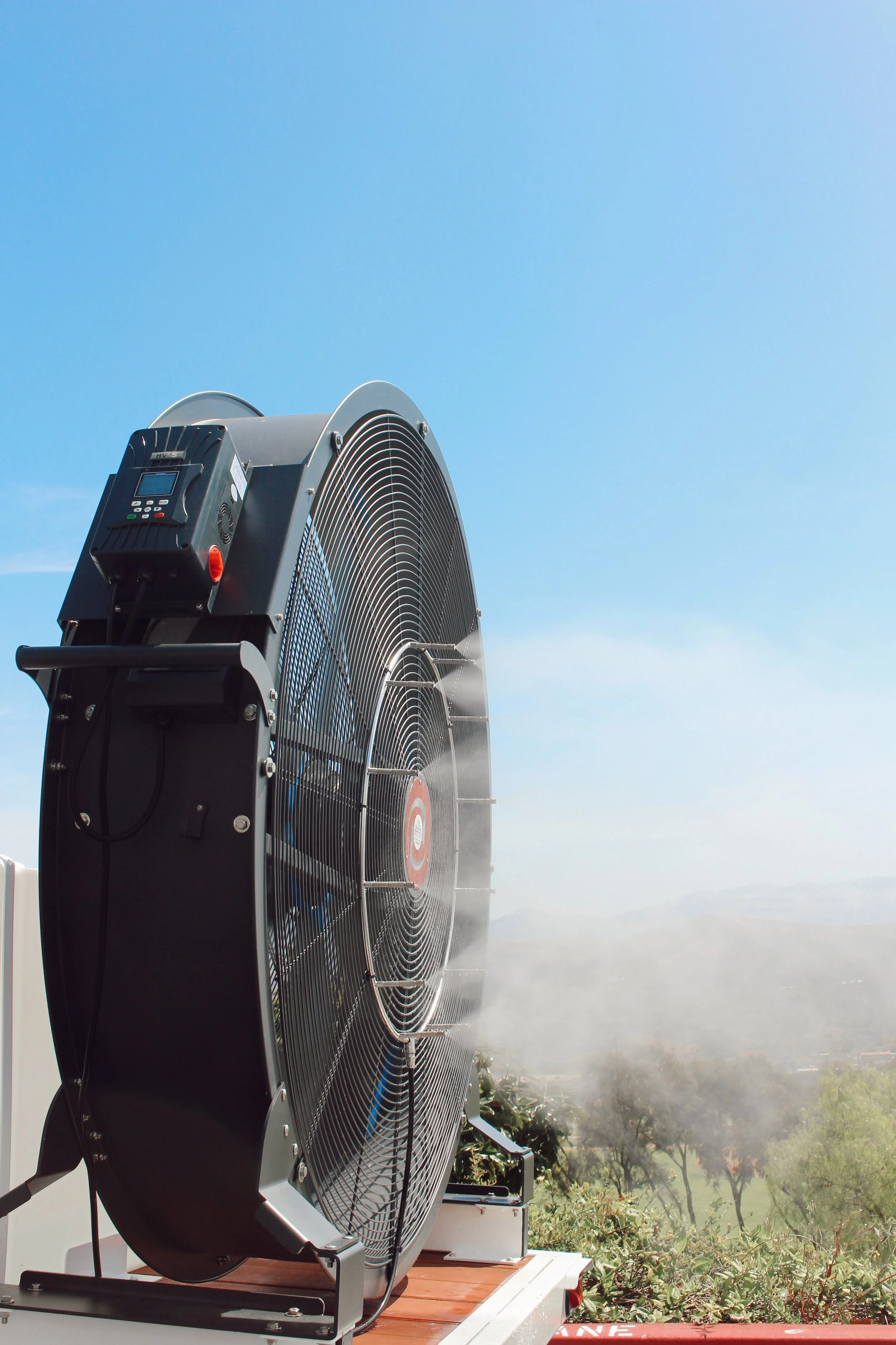 A large industrial cooling fan installed outdoors on a rooftop, with trees and a blue sky in the background.