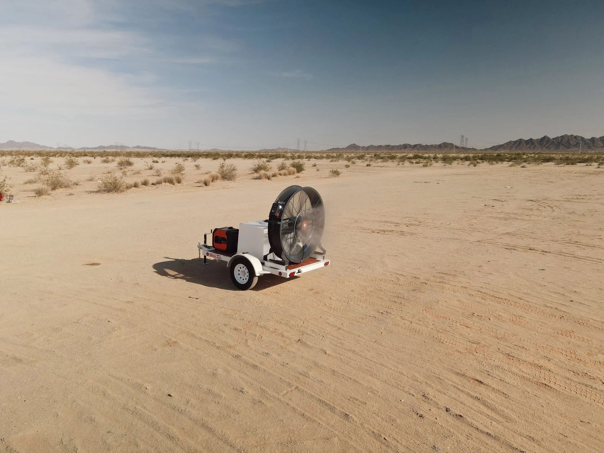 A desert landscape with sparse vegetation, distant mountains, and power lines under a cloudy sky. In the foreground, there is a portable misting fan on a small trailer.