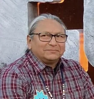 A man with gray hair and glasses wearing a red plaid shirt, standing outdoors near a gray wall with orange and black structure in the background.
