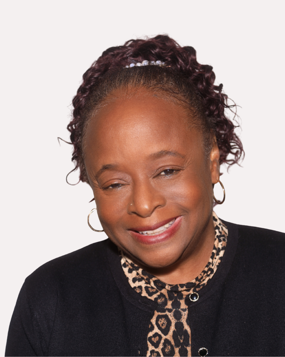 Close-up of a smiling African American woman with curly purple hair in a black top with leopard print details, wearing hoop earrings, against a plain white background.