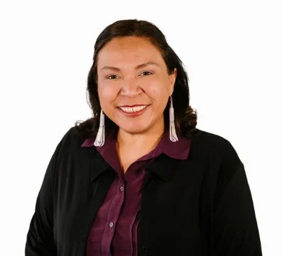 A woman with dark hair smiling, wearing earrings, a purple shirt, and a black jacket, posed against a white background.