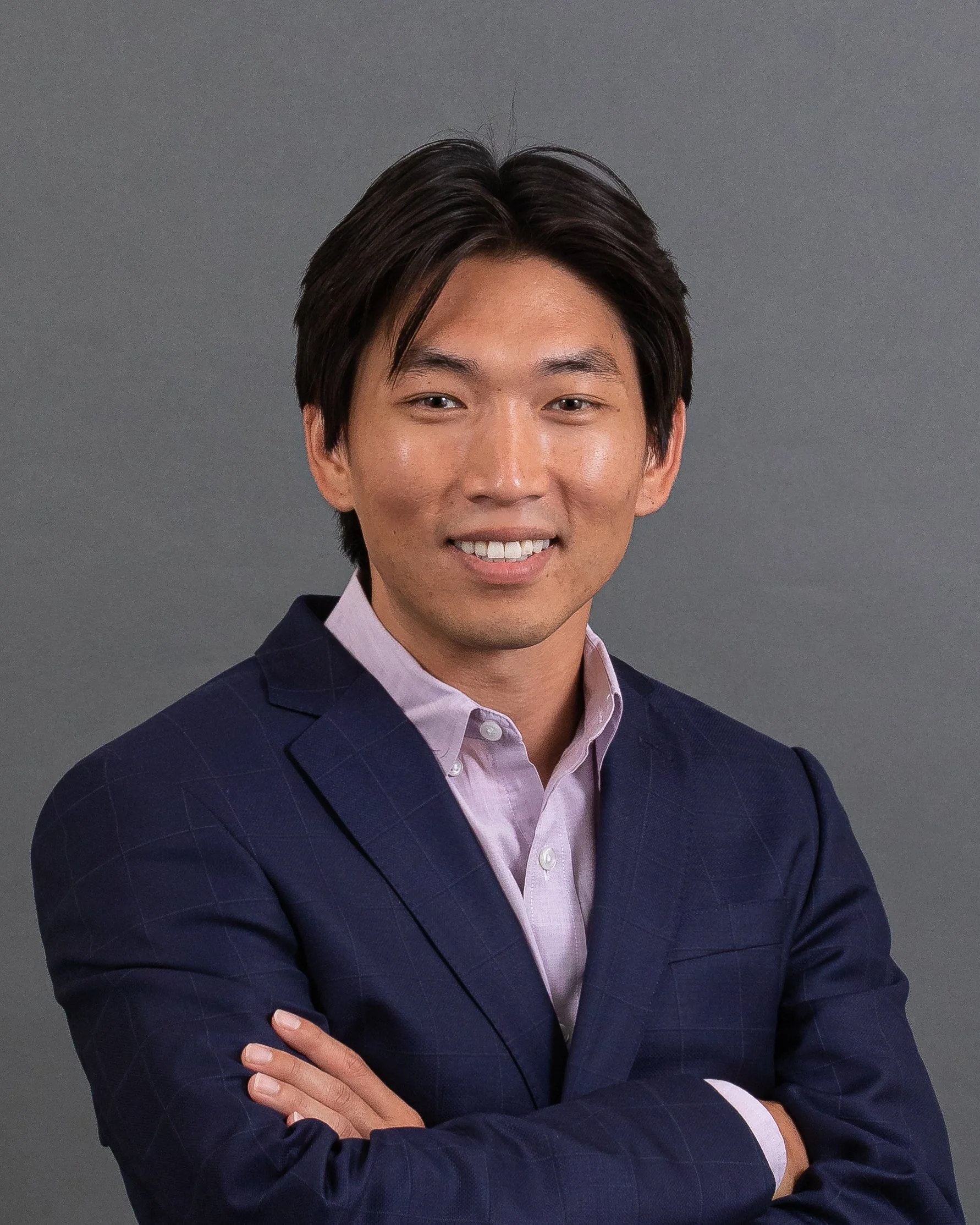 A young man with dark hair, wearing a dark checkered blazer and a light pink dress shirt, standing against a plain gray background, smiling with arms crossed.