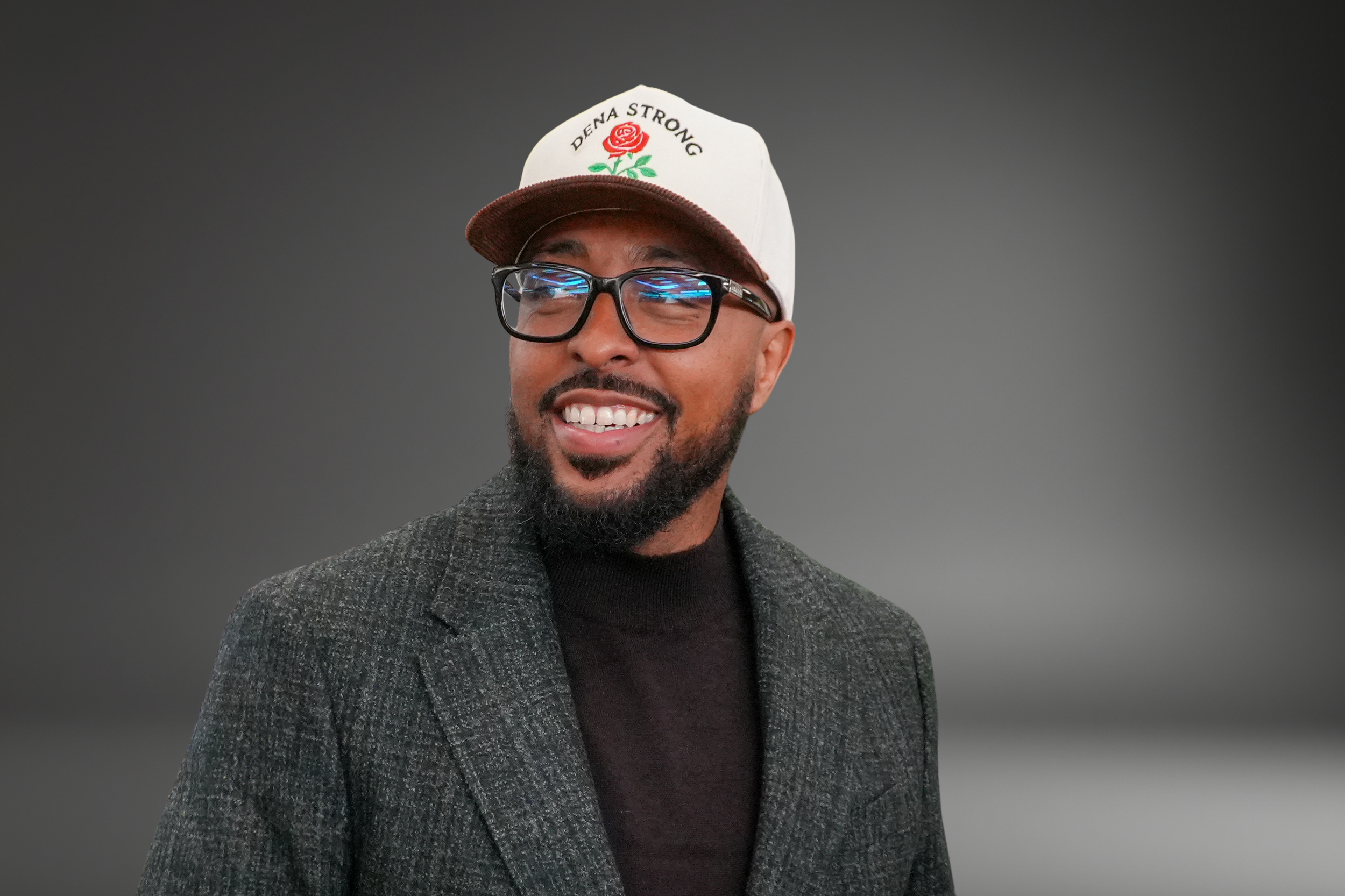 A smiling man wearing glasses, a gray blazer, dark shirt, and a white trucker hat with a red rose embroidered on the front and the words 'DENA STRONG' above it. He has a beard and dark hair.