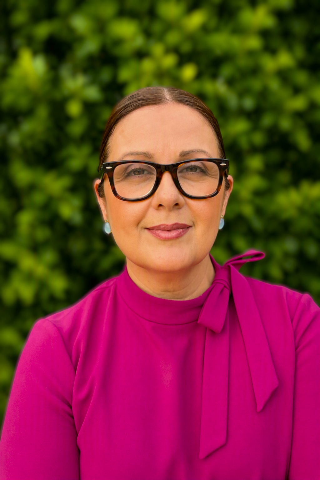 A woman with brown hair tied back, wearing large black glasses, a pink top with a bow on the shoulder, and earrings, standing outdoors with green foliage in the background.
