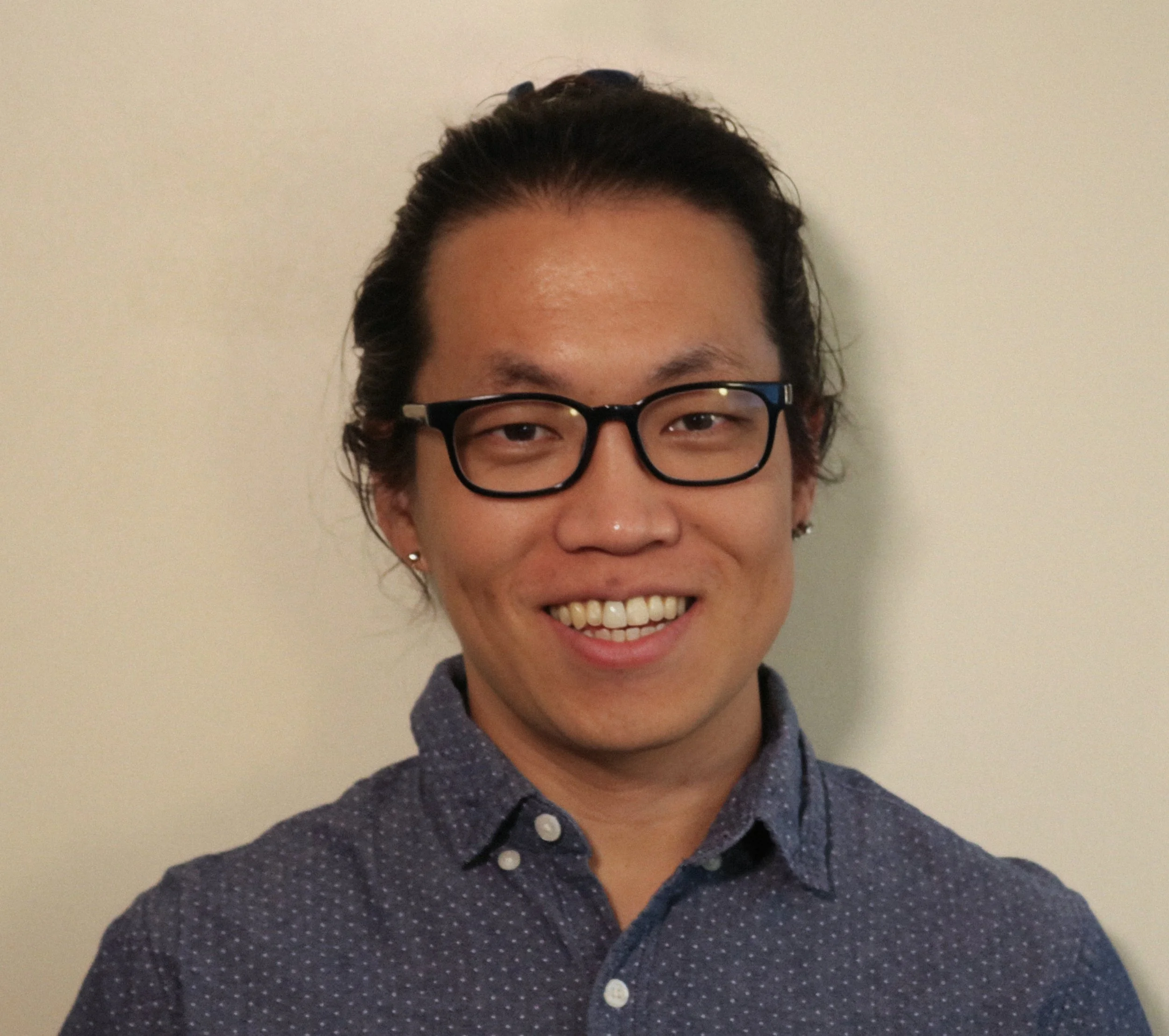 A person with glasses smiling, wearing a dark, patterned button-up shirt, against a plain light-colored wall.