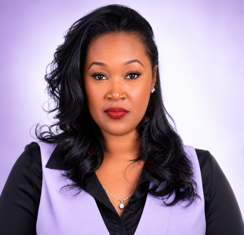A woman with long, wavy black hair, wearing makeup including red lipstick, and dressed in a black top with a lavender vest and a pearl necklace, against a light purple background.