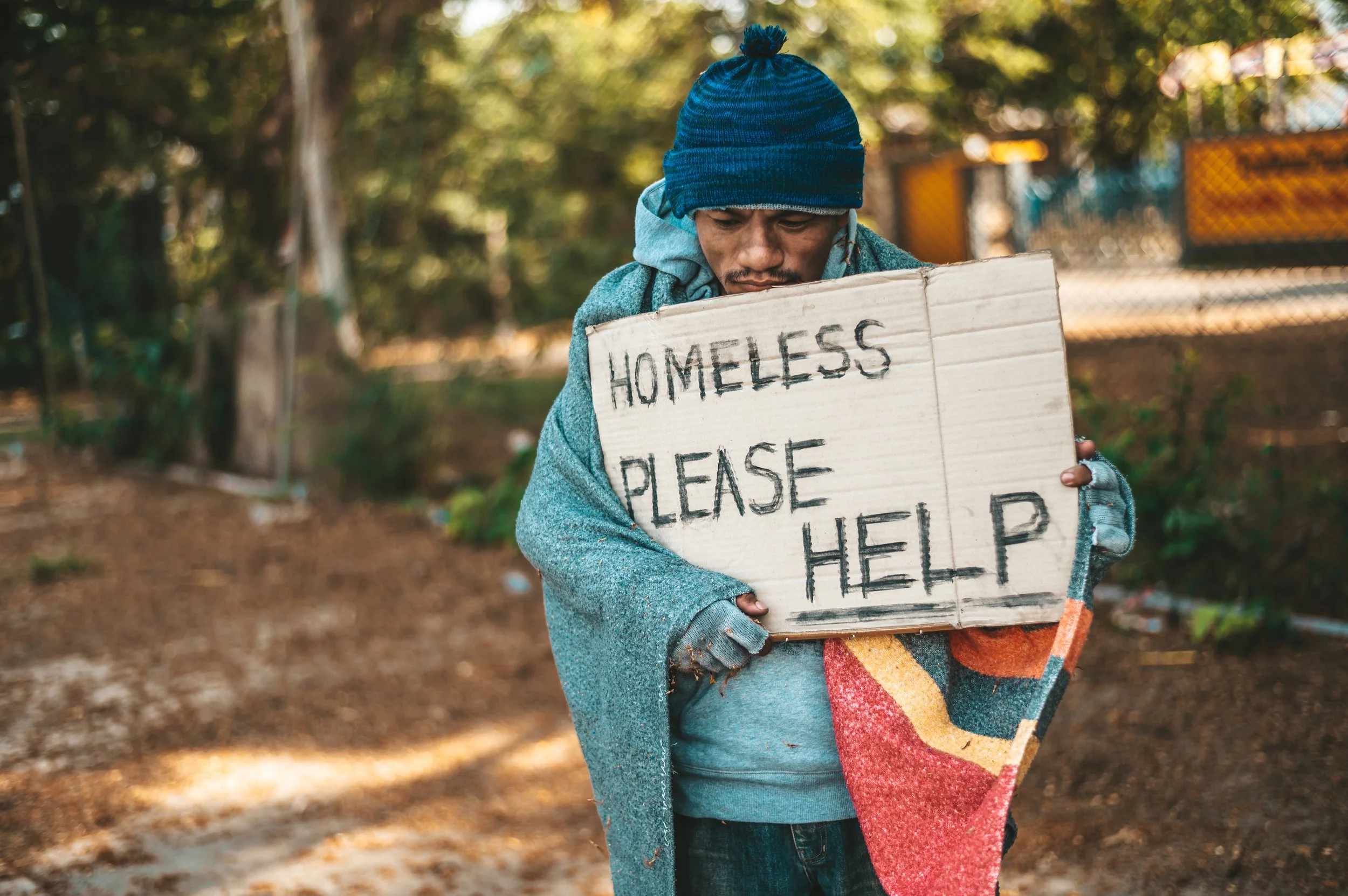 Man wearing a blue beanie and gray sweater holding a cardboard sign that reads 'Homeless Please Help' in an outdoor setting with trees and a fence in the background.