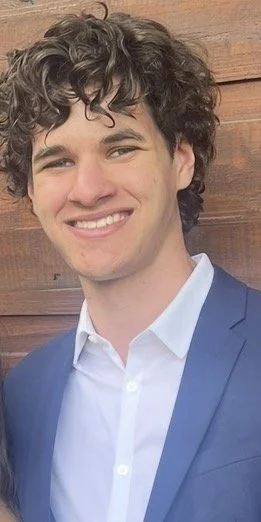 Young man with curly hair smiling in front of a wooden background, wearing a blue blazer and white shirt.