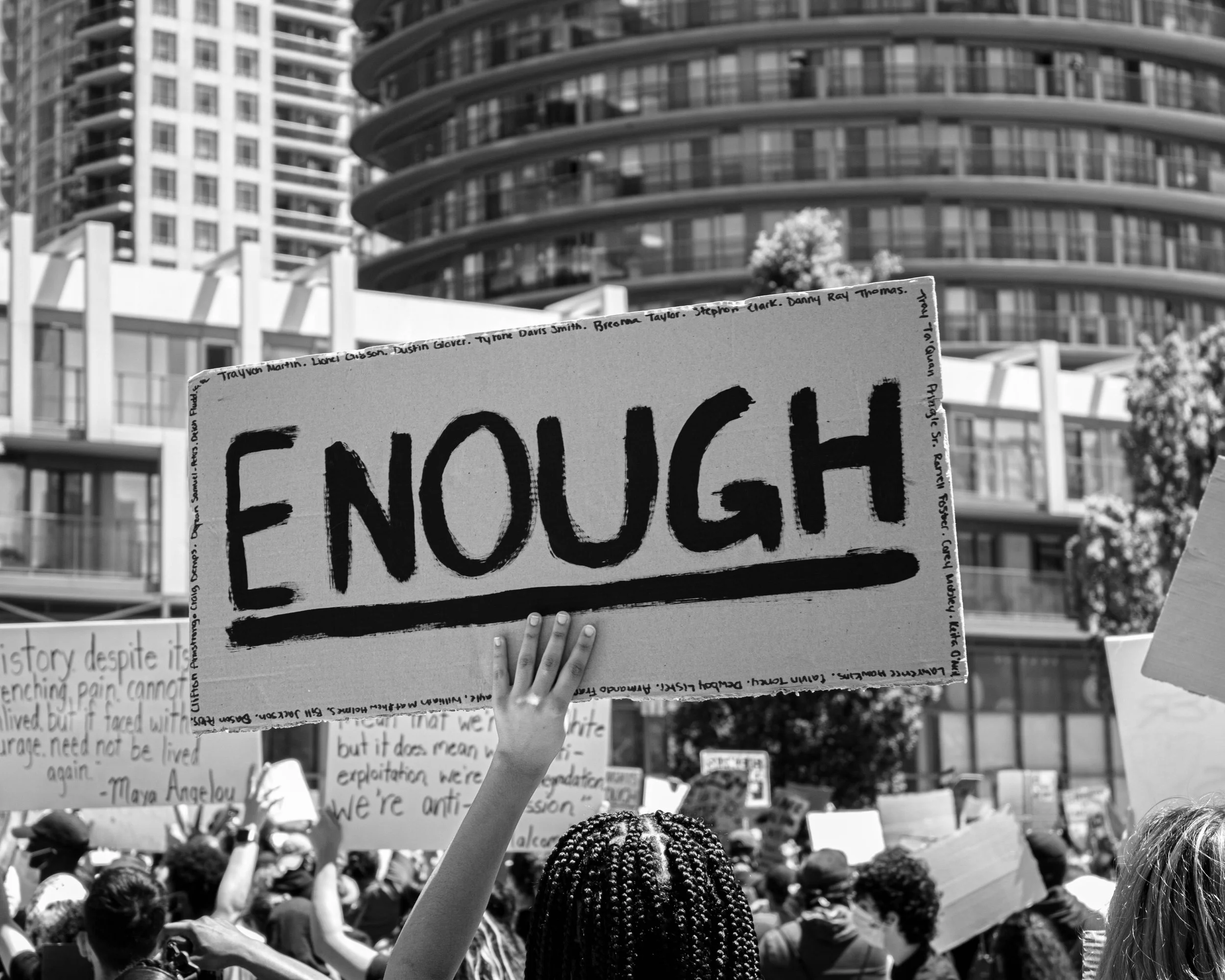 Black and white image of a protest. A woman's hand holds up a sign with the word "Enough".
