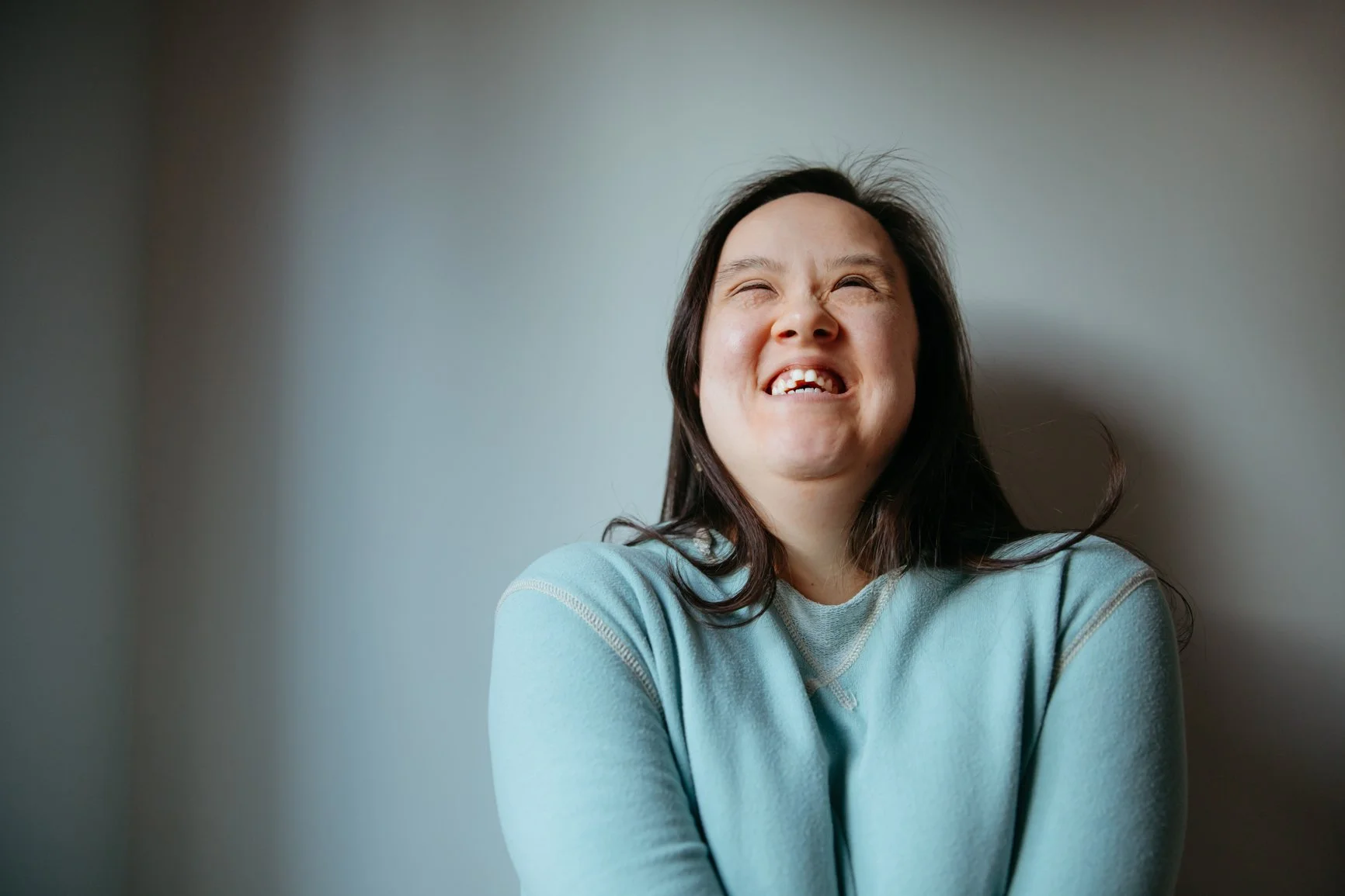 Photo of a young disabled woman with long brown hair looking up and laughing. She is wearing a light blue top and is in front of a grey background.