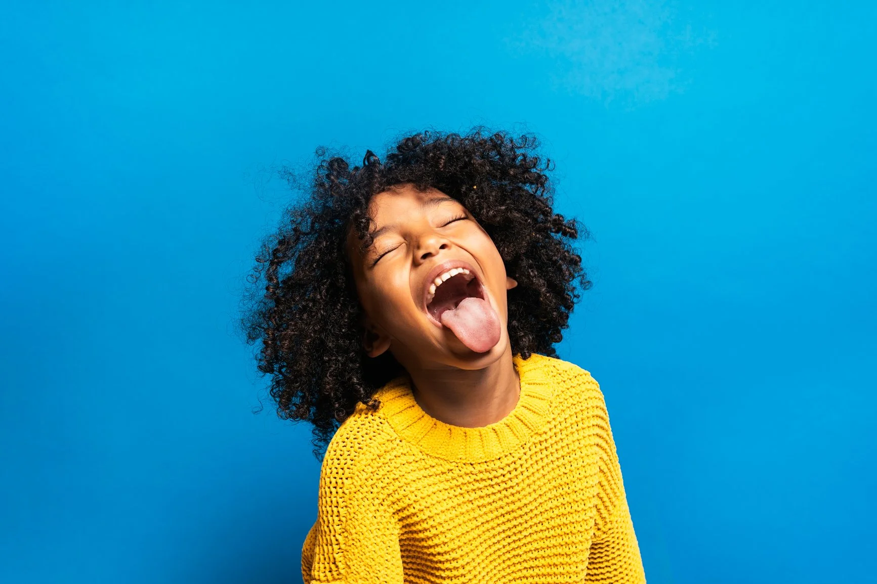 Image of a young Black or mixed race girl with curly hair against a blue background. She is wearing a yellow sweatshirt and sticking her tongue out.