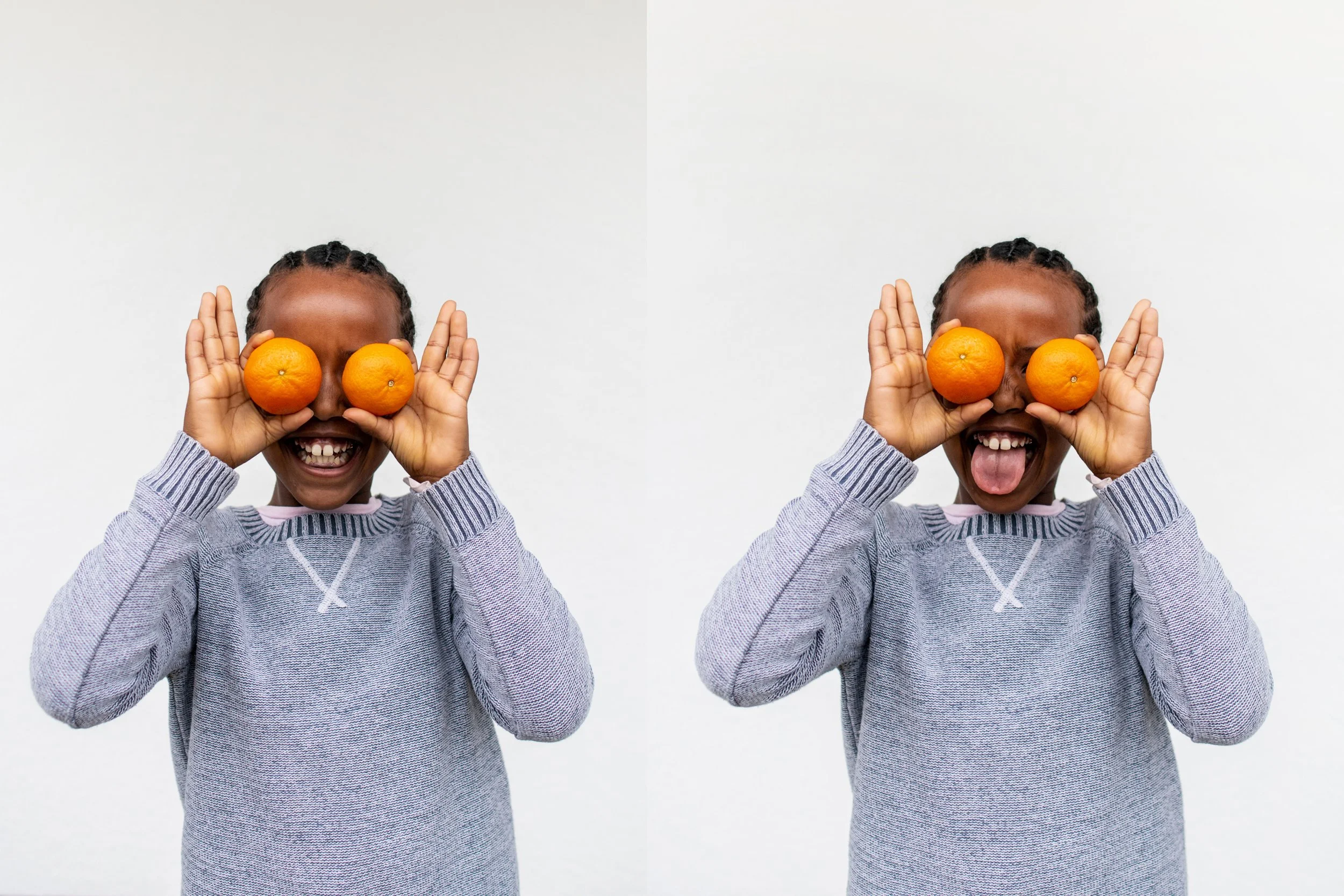 Two Black girls in cornrows and a grey sweater. They are holding oranges in in front of their eyes. One is smiling and the other has their tongue sticking out.
