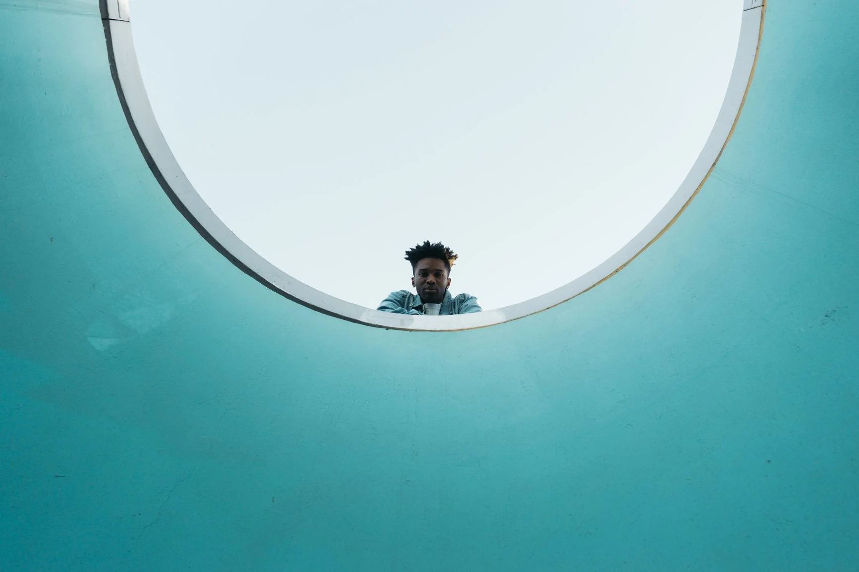 A young Black man with short dreads is looking down over the edge of an agua blue circular structure. He is wearing a white T-shirt under a blue shirt.