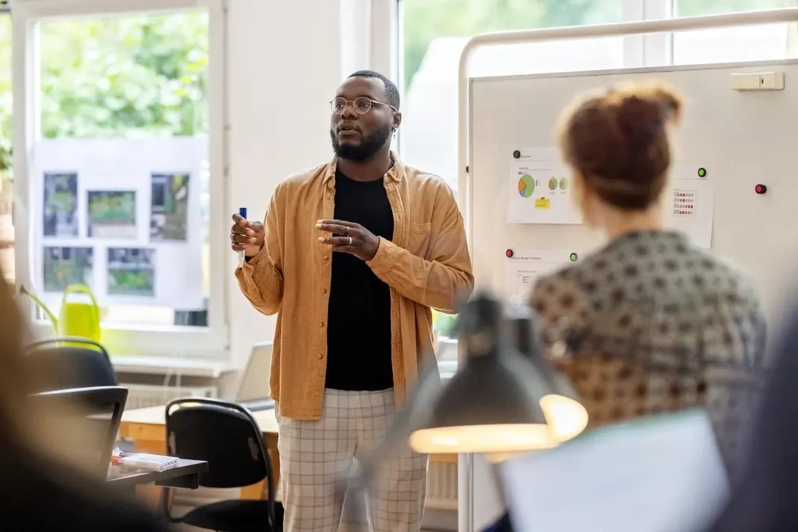 Black man with glasses is presenting in an office. He is dressed in a black t-shirt with an open orange shirt on top. He holds a marker in front of a bulletin board.