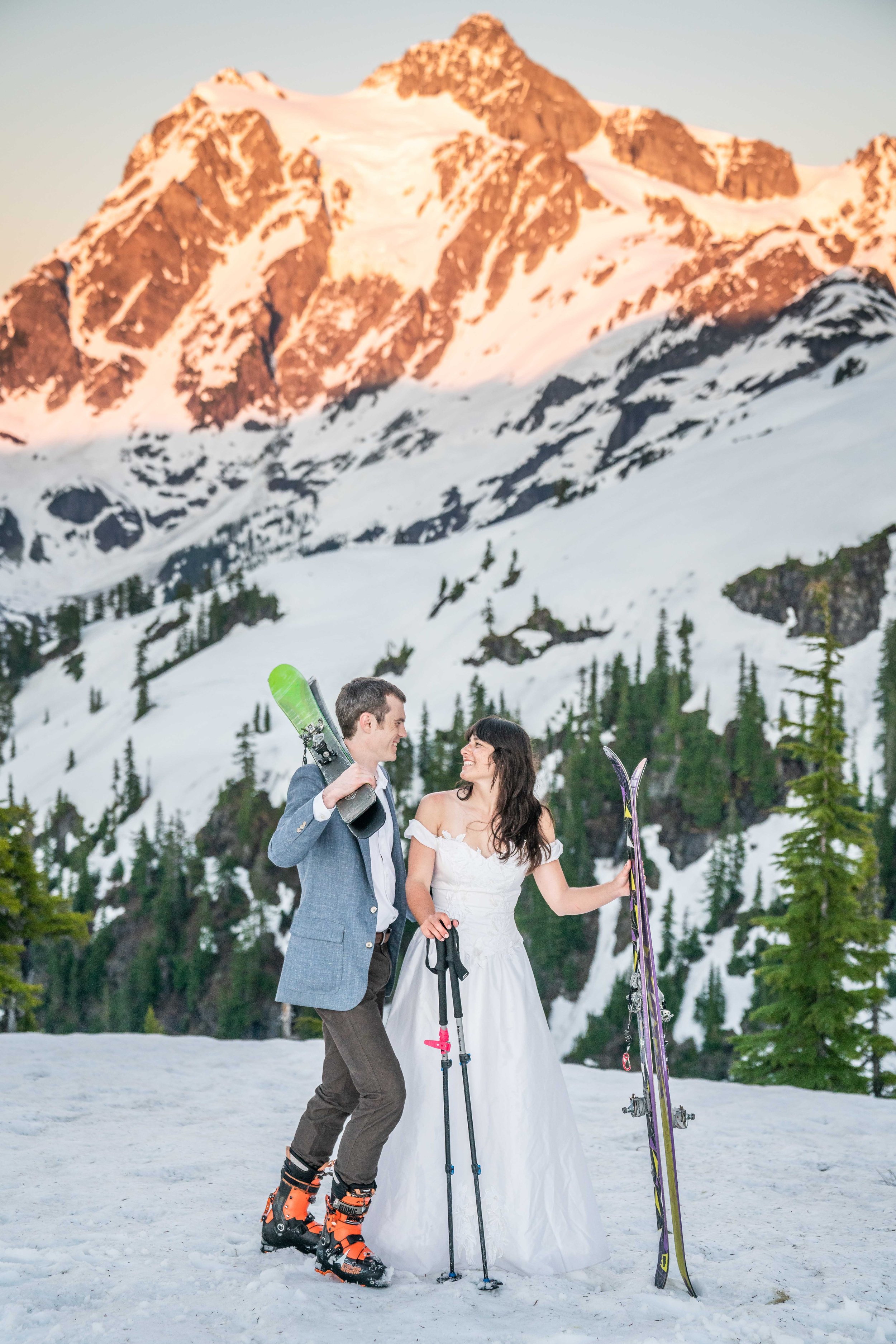 A photo of a couple in wedding clothes standing together in the snow. The man is wearing ski boots and both are holding skis. Mount Shuksan at sunset is behind them.