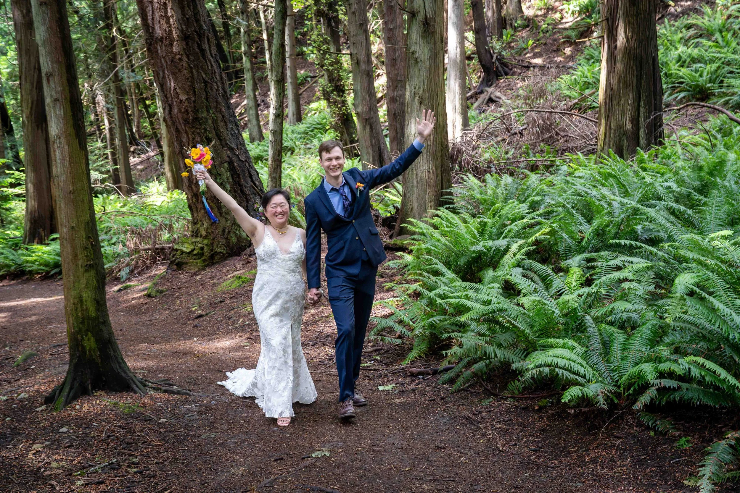 A bride and groom hold hands and walk towards the camera on the Fragrance Lake Trail in Larrabee State Park shortly after eloping. They have their hands raised in celebration and they are surrounded by lush ferns and old growth trees.