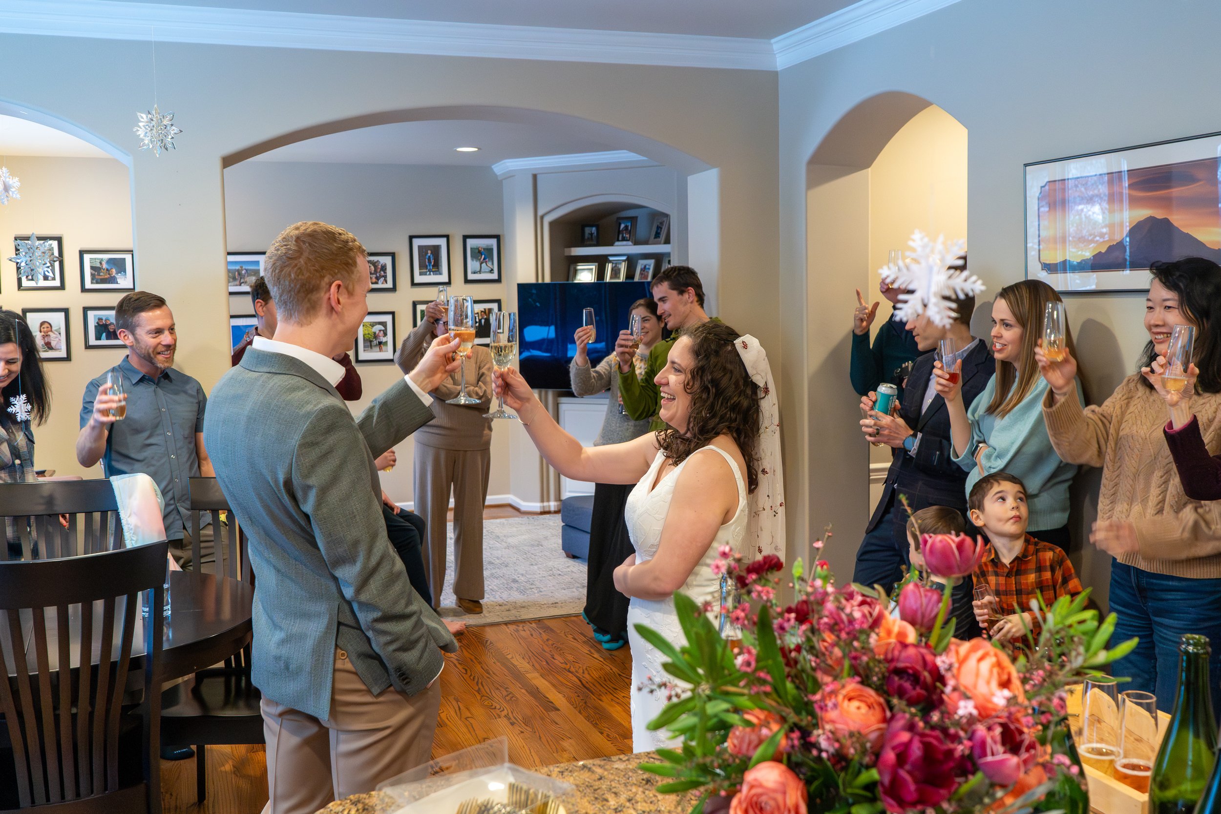 Photo of a just-married couple in wedding clothes raising champagne glasses in a toast. They're in a brightly lit home surrounded by friends and family who are also raising glasses.