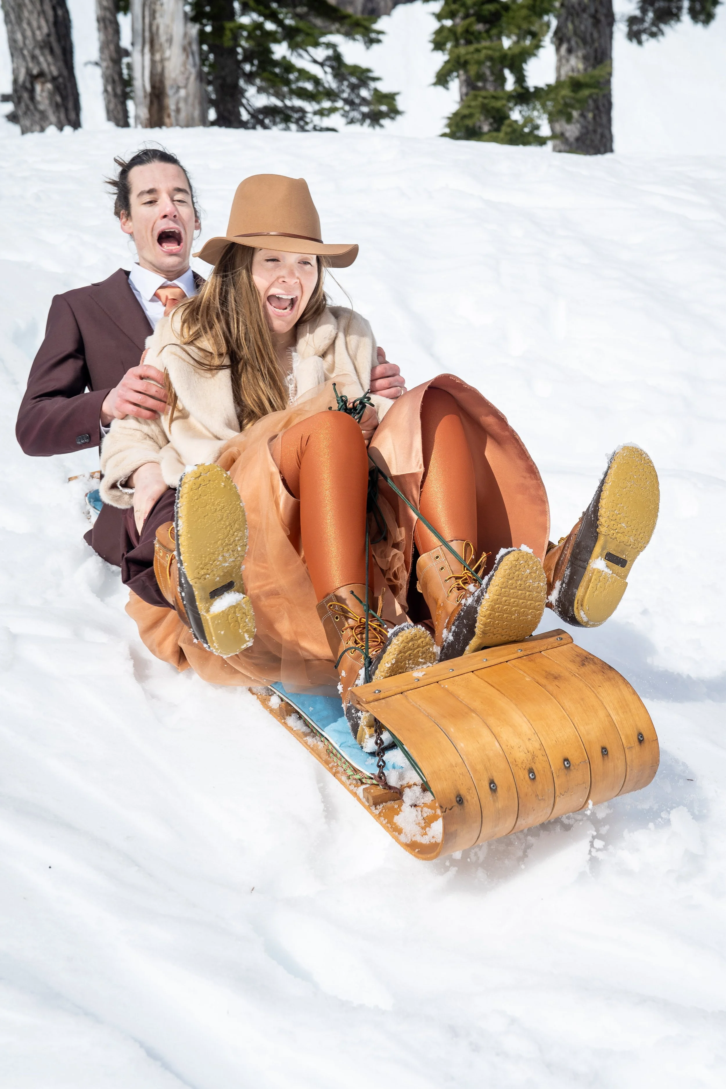 A bride and groom in wedding clothes yelling as they ride downhill on snow on a wooden toboggan after getting married.