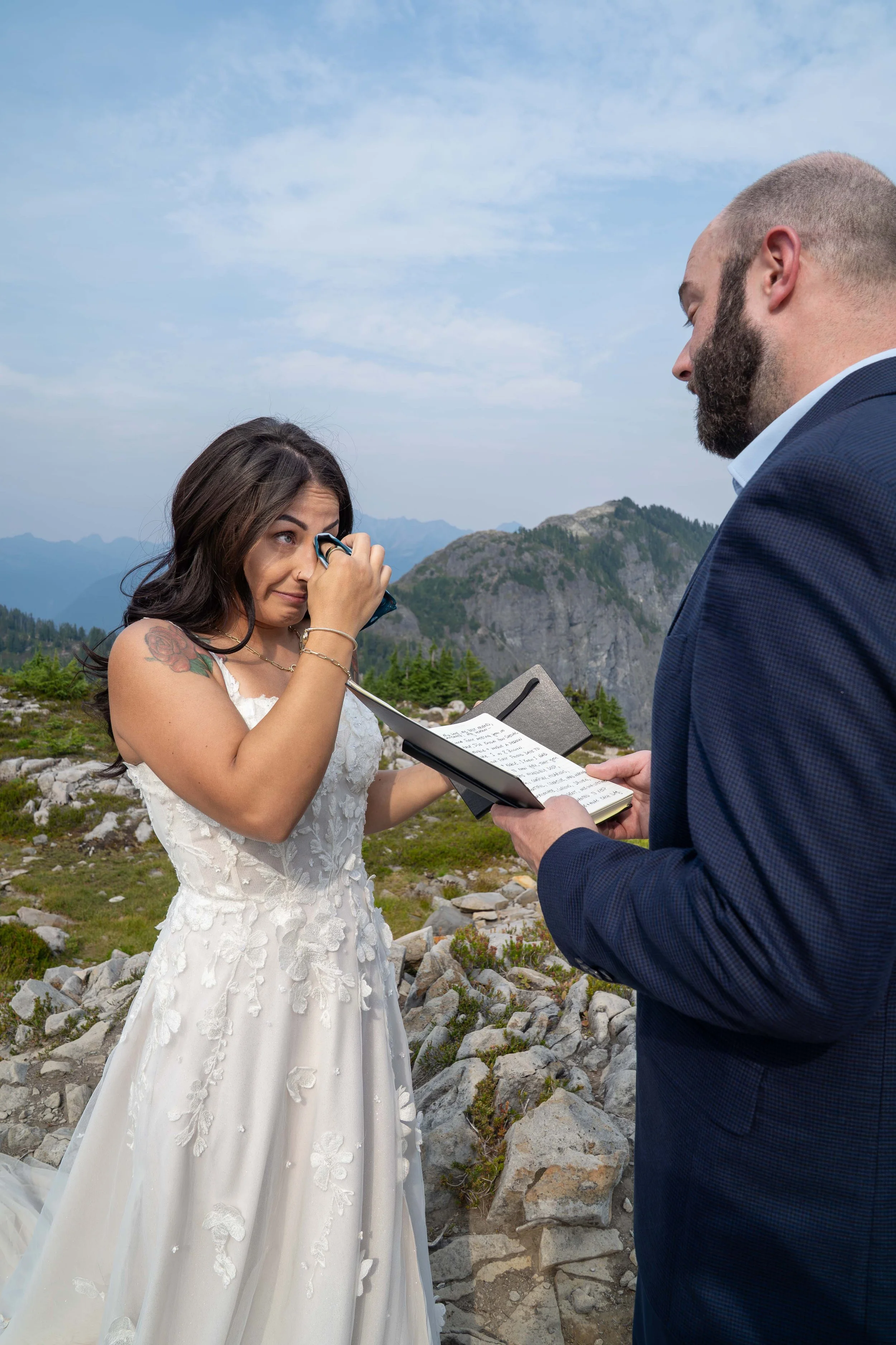 A groom reading his handwritten vows from a vow book during an elopement ceremony at Artist Point, near Mt. Baker, while his bride wipes tears from her eyes. They are standing in a rocky alpine meadow with the North Cascades behind.