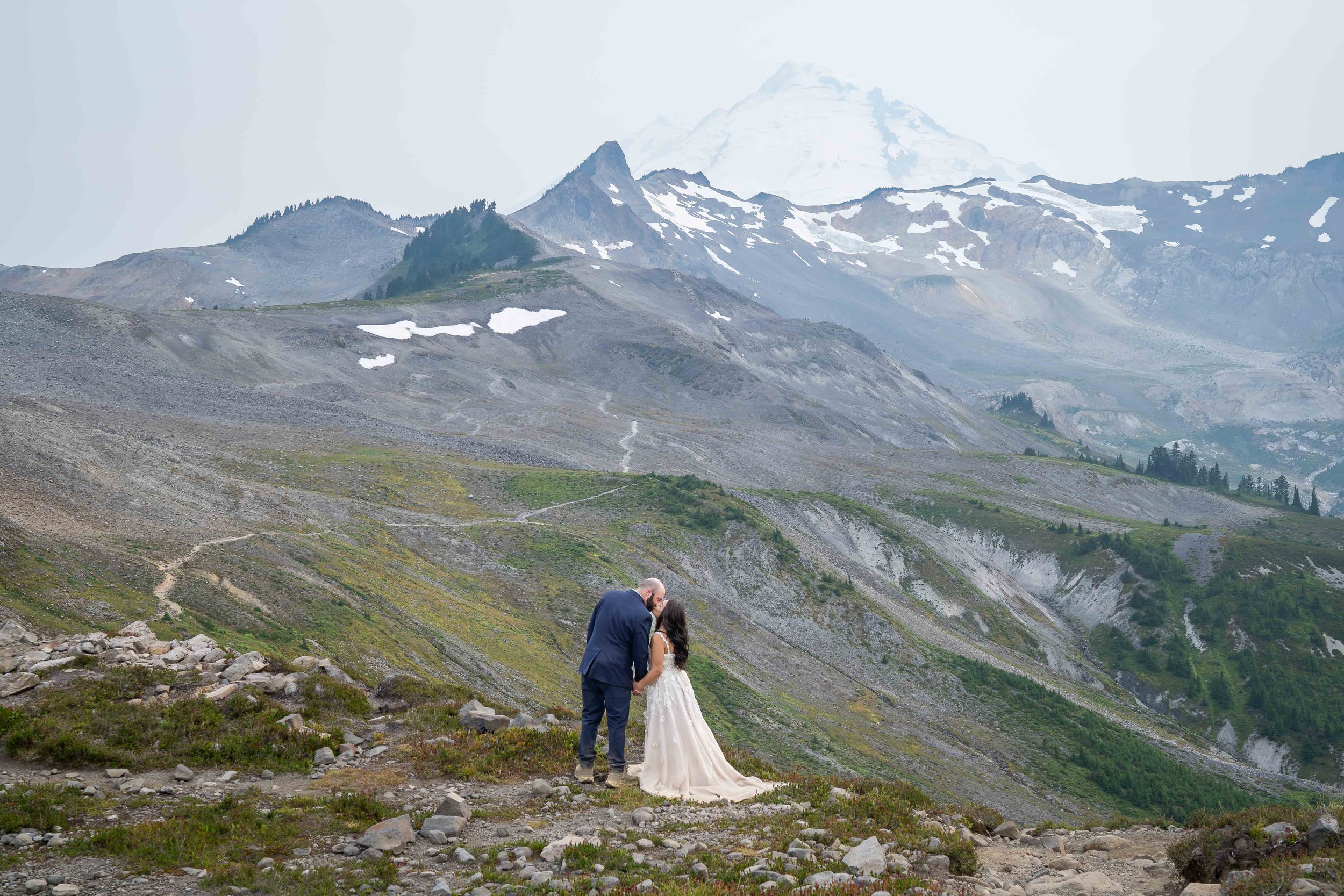 A bride and groom kiss in front of Mt. Baker at sunset during their Ptarmigan Ridge elopement. The photo is wide-angle, showing the scale of the landscape and the Ptarmigan Ridge trail stretching towards Coleman Pinnacle and Mt. Baker.