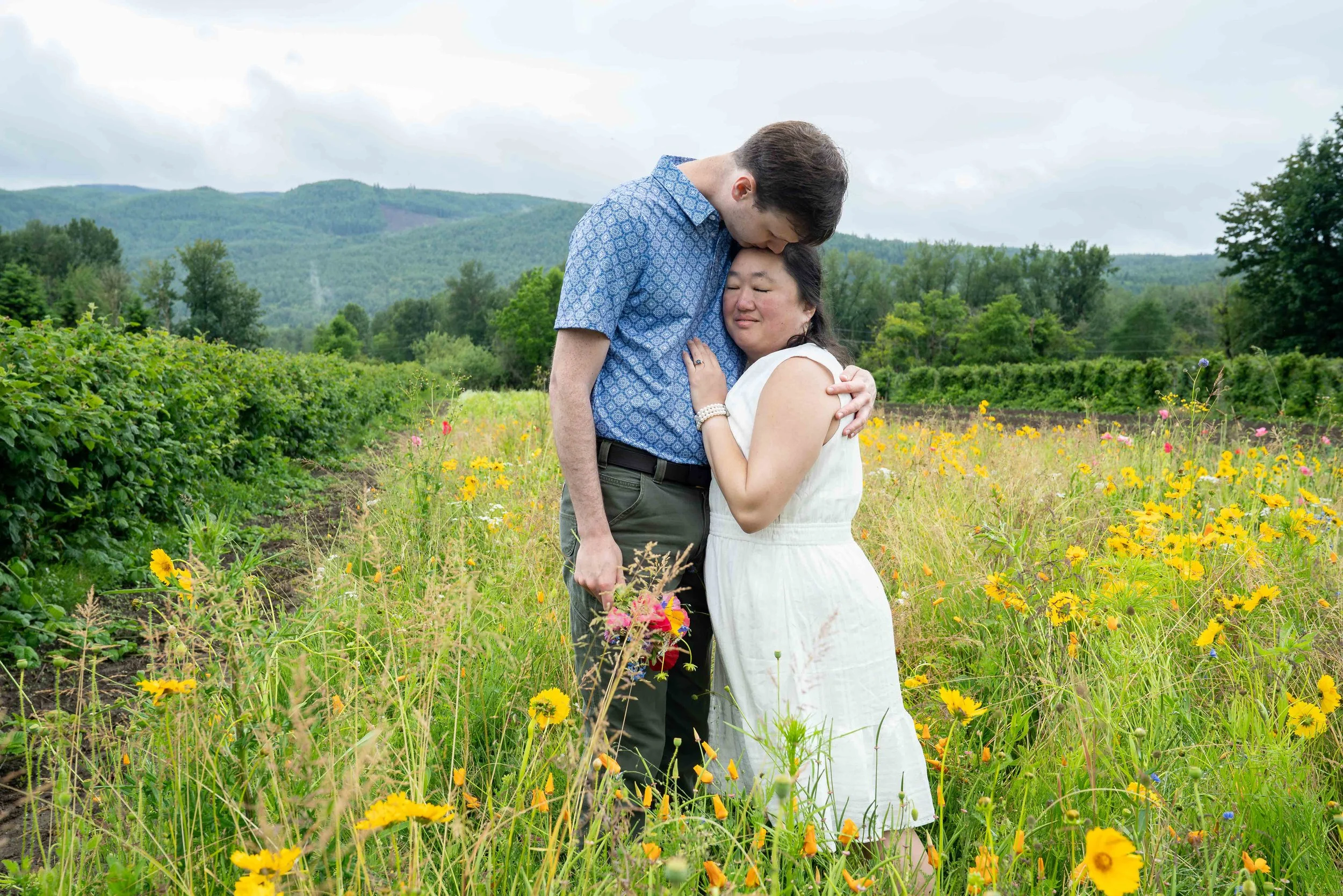 A bride and groom standing together in a field of yellow flowers during a Bellingham elopement photography session. The bride's head is pressed against the groom's chest and he is leaning down to kiss her.
