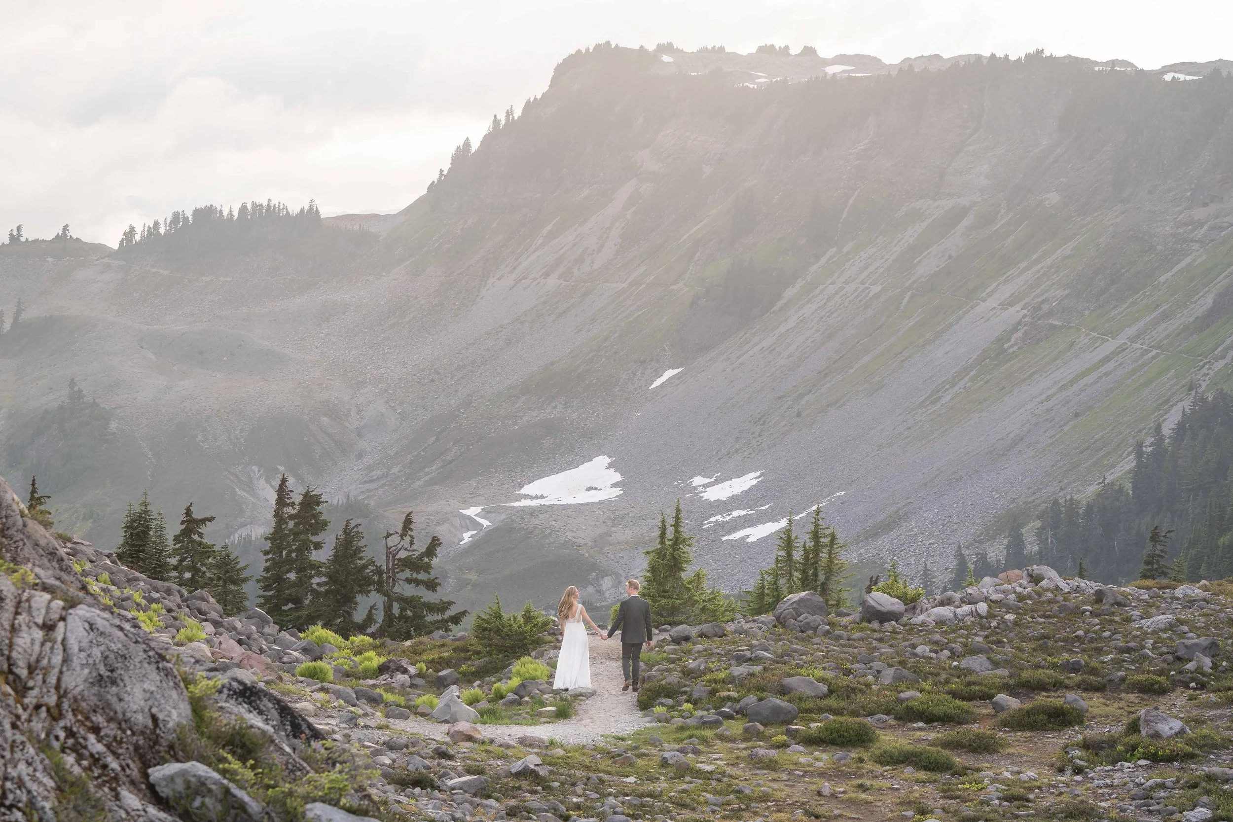 Photo of a bride in a wedding dress and a groom in a suit walking on a trail at Artist Point, Washington, at sunset after eloping. The couple is walking away from the camera towards Table Mountain, which is clearly visible in front of them.