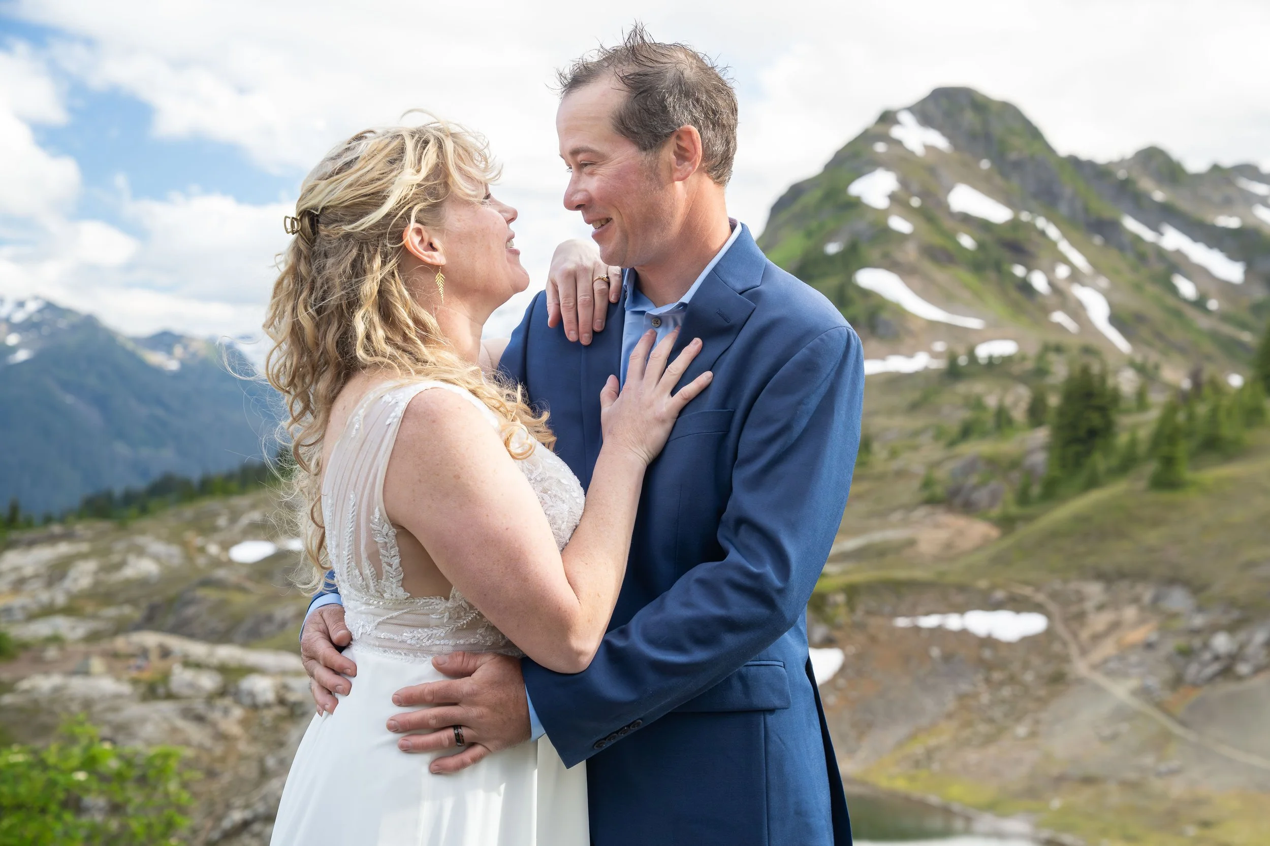 A bride and groom in wedding clothes holding each other closely during their North Cascades elopement near Yellow Aster Butte. Subalpine meadows and snowy peaks are visible in the background.