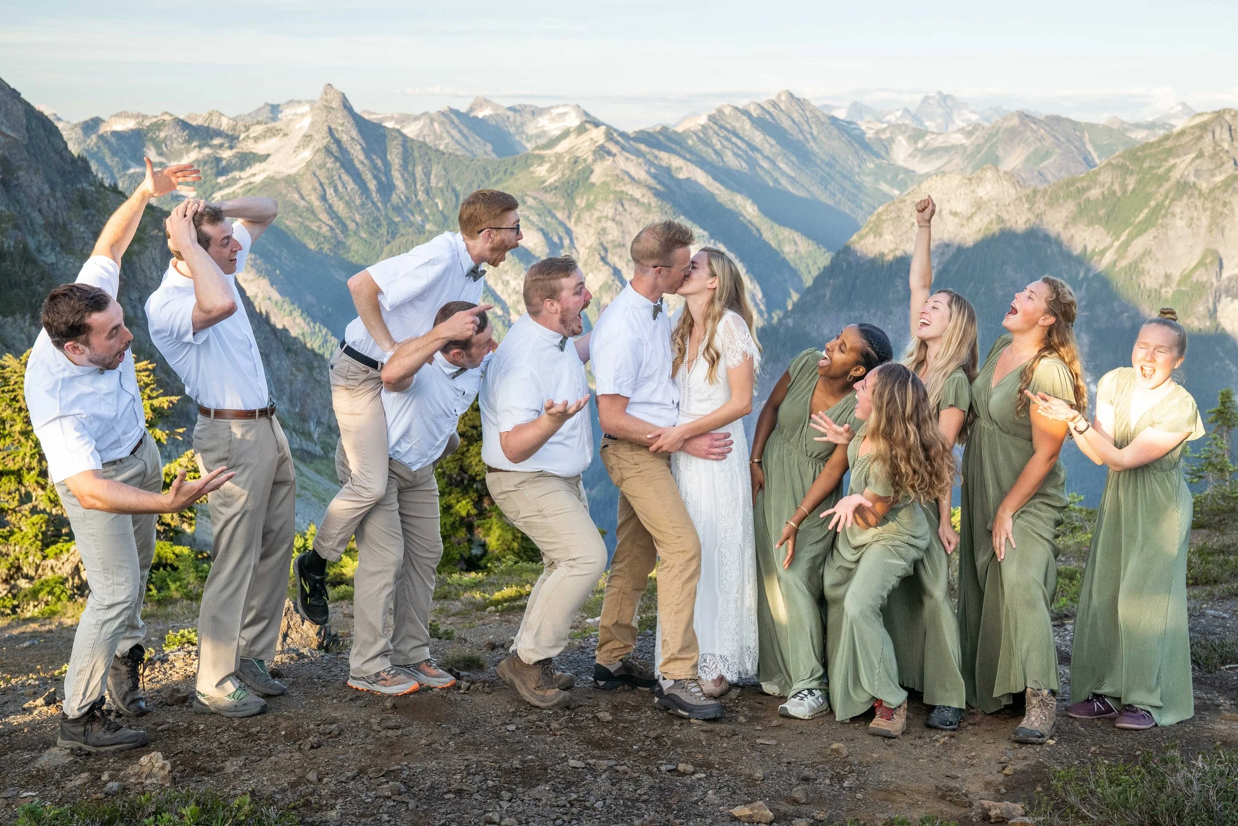 A group photo of a wedding party during a North Cascades elopement. The bridesmaids and groomsmen are acting shocked to see the couple kiss and mountains are visible behind them.