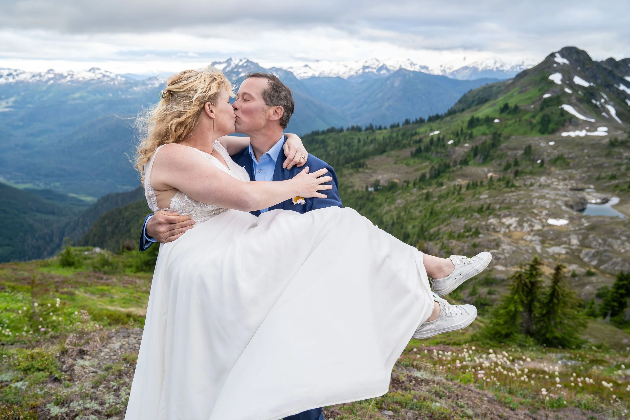 A groom in a suit holding a bride in a wedding dress and kissing during a North Cascades elopement at Yellow Aster Butte, in Washington. They are standing in a subalpine meadow of wildflowers with an alpine lake and Mt. Baker visible behind them.
