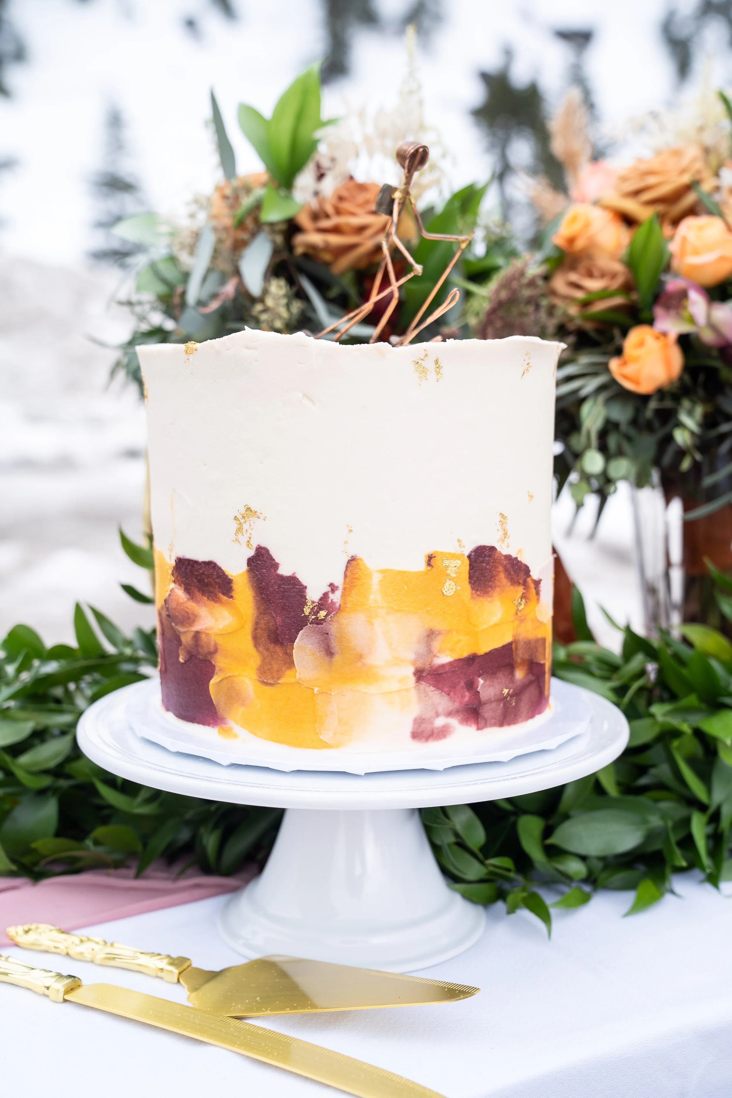 A photo of a wedding cake on a tray. The cake is white with orange and red trim, and behind is is a bouquet of green leaves and orange roses.