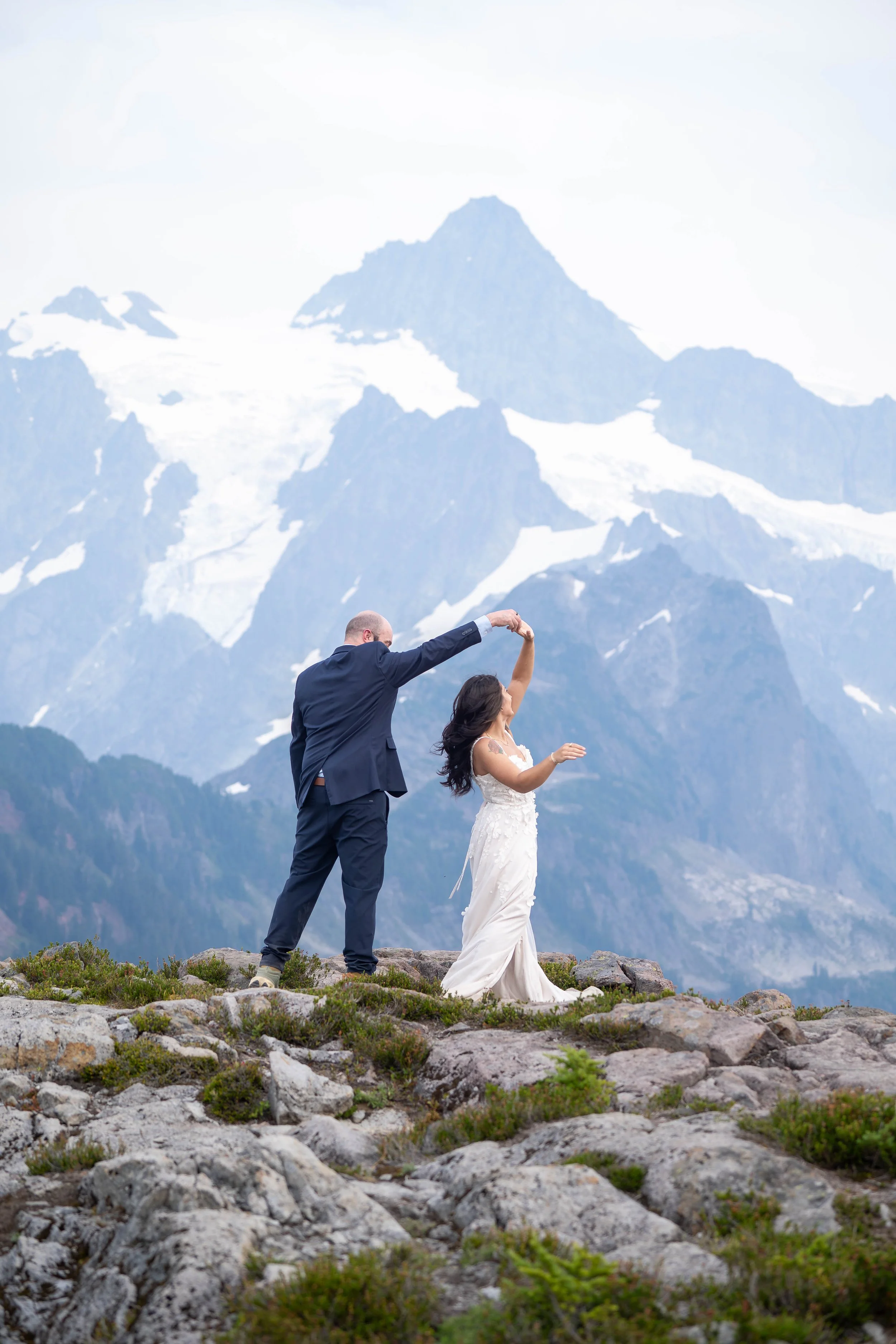 A groom spinning his bride by the hand in front of Mt. Shuksan during an Artist Point elopement. They are dancing in a rocky alpine meadow with the glacier-covered mountain looking huge behind them.